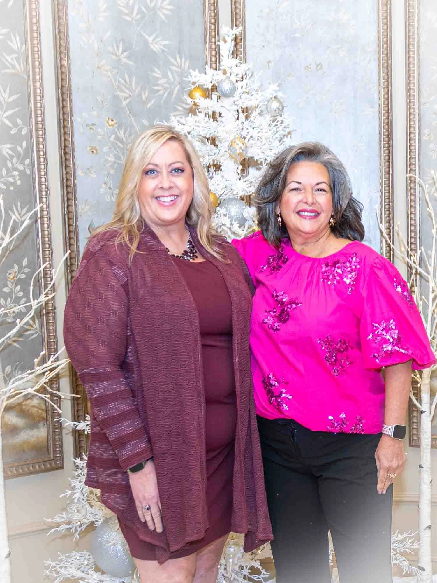 Two women smiling in front of a white Christmas tree with ornaments, framed background art visible.