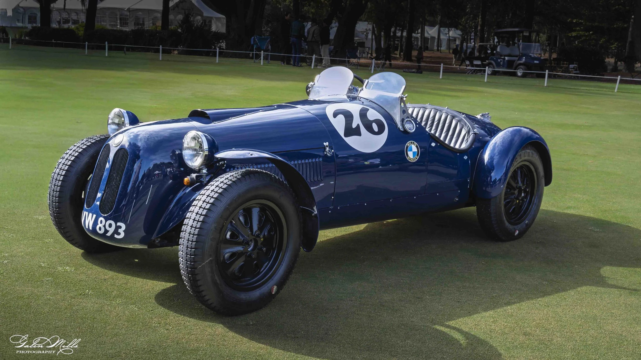 A vintage blue race car with the number 26 on the side, parked on a grassy area at a car show or event, with several people and golf carts in the background.