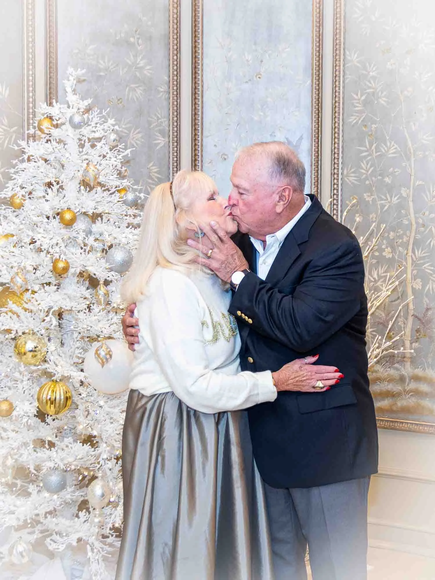 Elderly couple kissing in front of white Christmas tree with gold and silver ornaments.