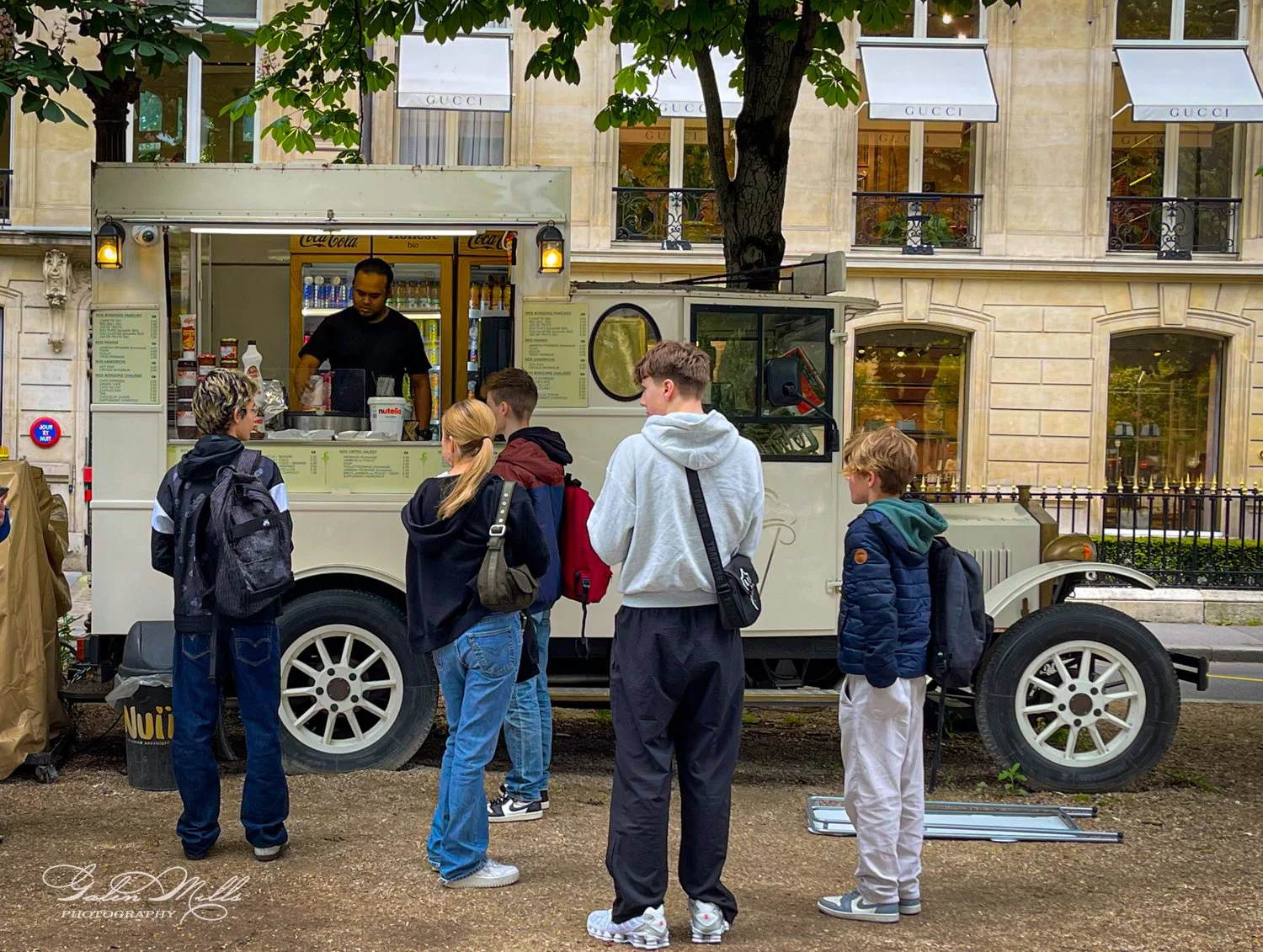 People standing in line at a street food truck with a vendor preparing food, parked near a building with Gucci store signage.