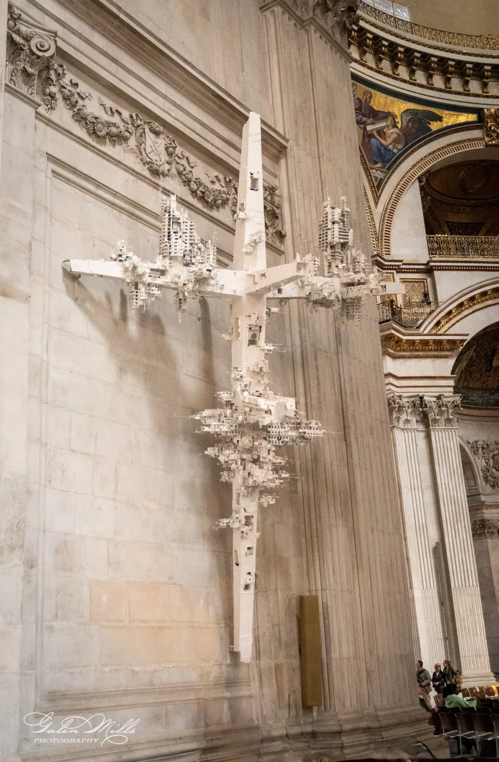 Art installation of a large white abstract sculpture resembling a cross with intricate details, mounted on a wall inside a historic building with ornate decor and murals.