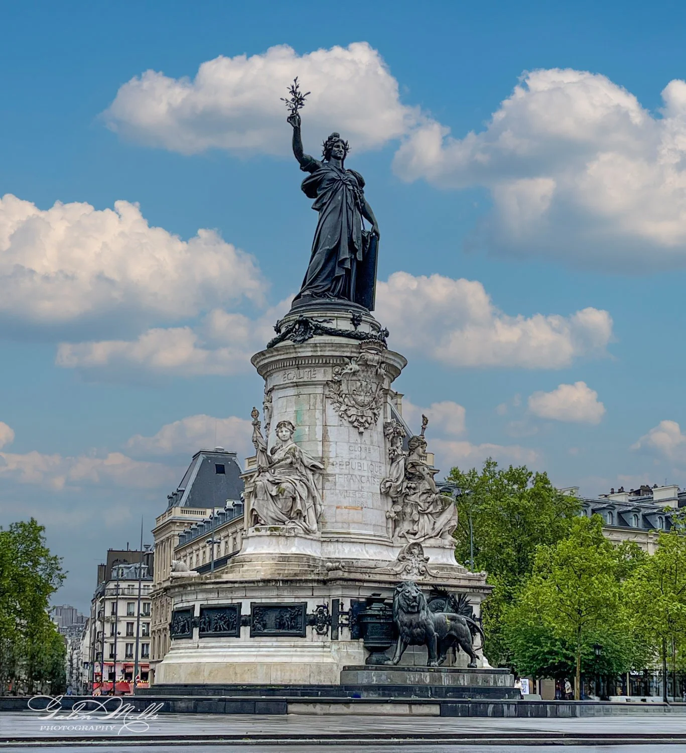 Statue of Marianne at Place de la République in Paris, surrounded by intricate sculptures, with a clear blue sky and clouds in the background.