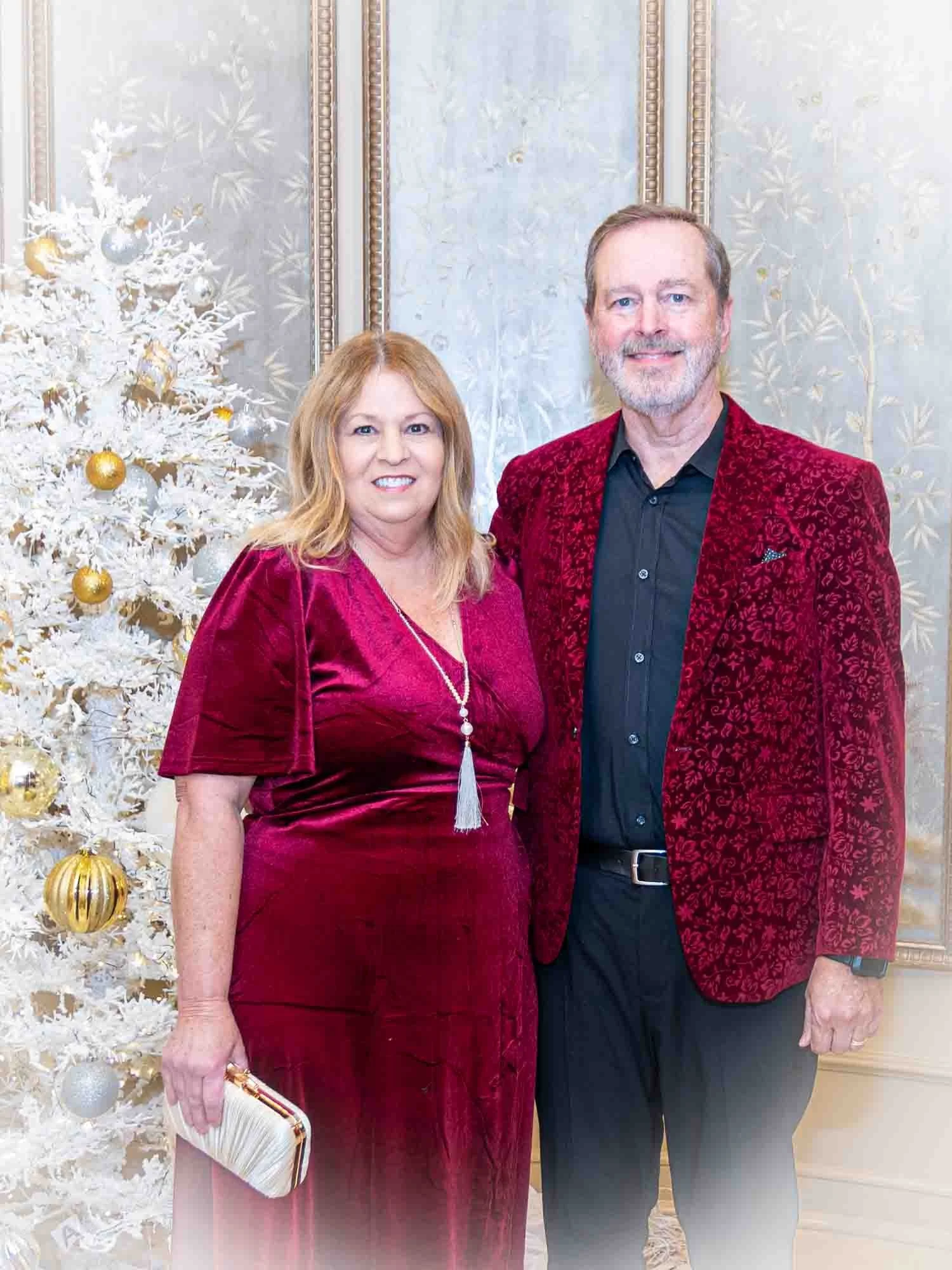 A man and woman wearing matching red formal attire standing next to a white Christmas tree decorated with gold and silver ornaments.