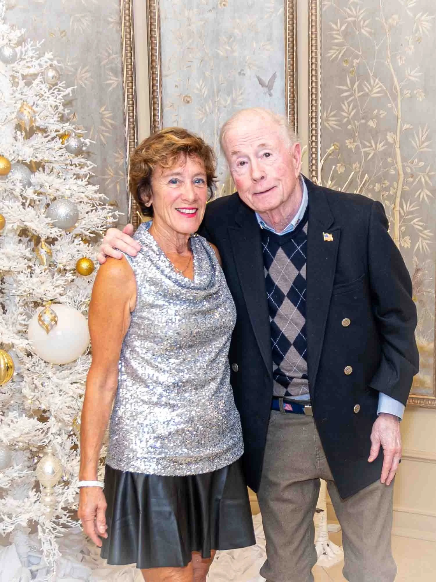 A smiling woman in a silver dress and an older man in a blazer stand beside a white Christmas tree decorated with gold and silver ornaments. They are positioned in front of an ornate wall with floral designs.