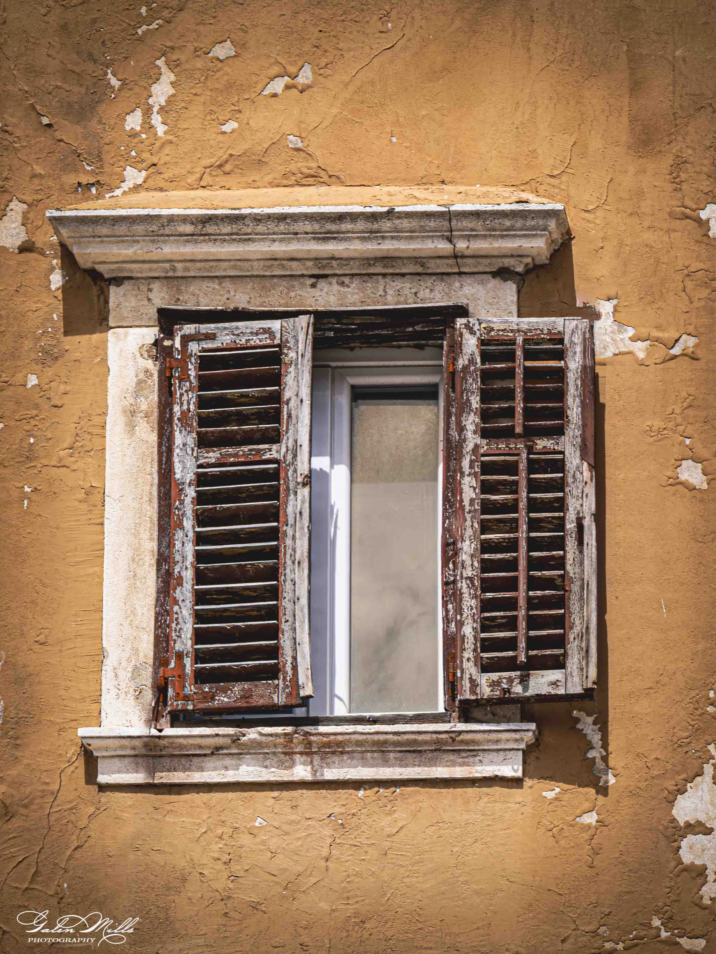 An old wooden window with shutters on an orange, weathered wall with peeling paint.