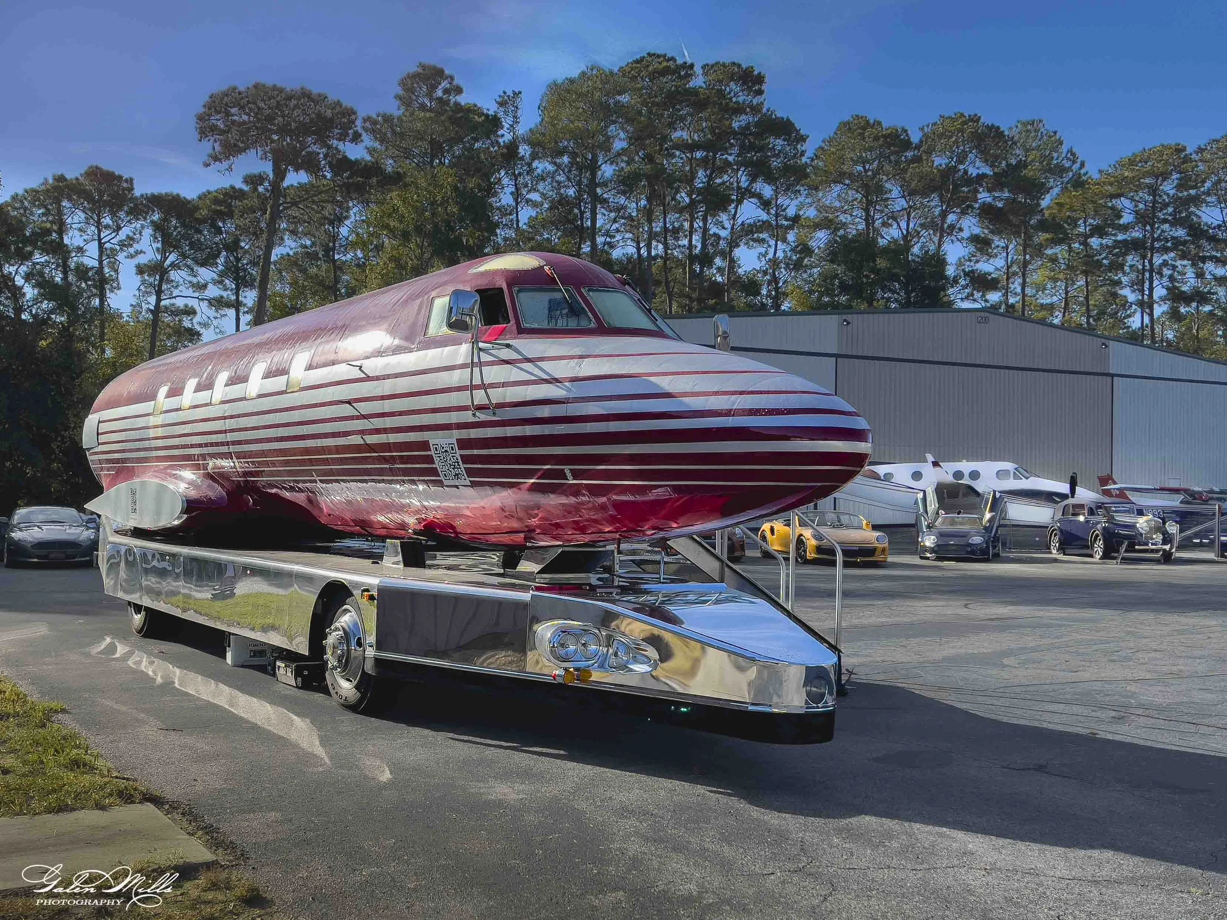 A red and white spaceship model displayed on a trailer in a parking lot with other cars and an airplane in the background.