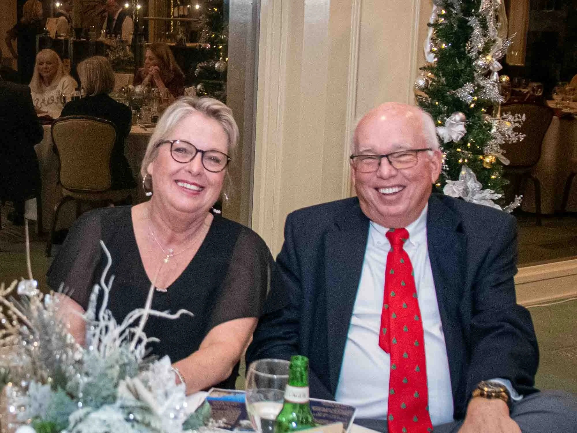 A smiling man and woman sitting at a decorated table at a festive event. The man is wearing a suit with a red tie, and the woman is wearing a black top. A Christmas tree is visible in the background.