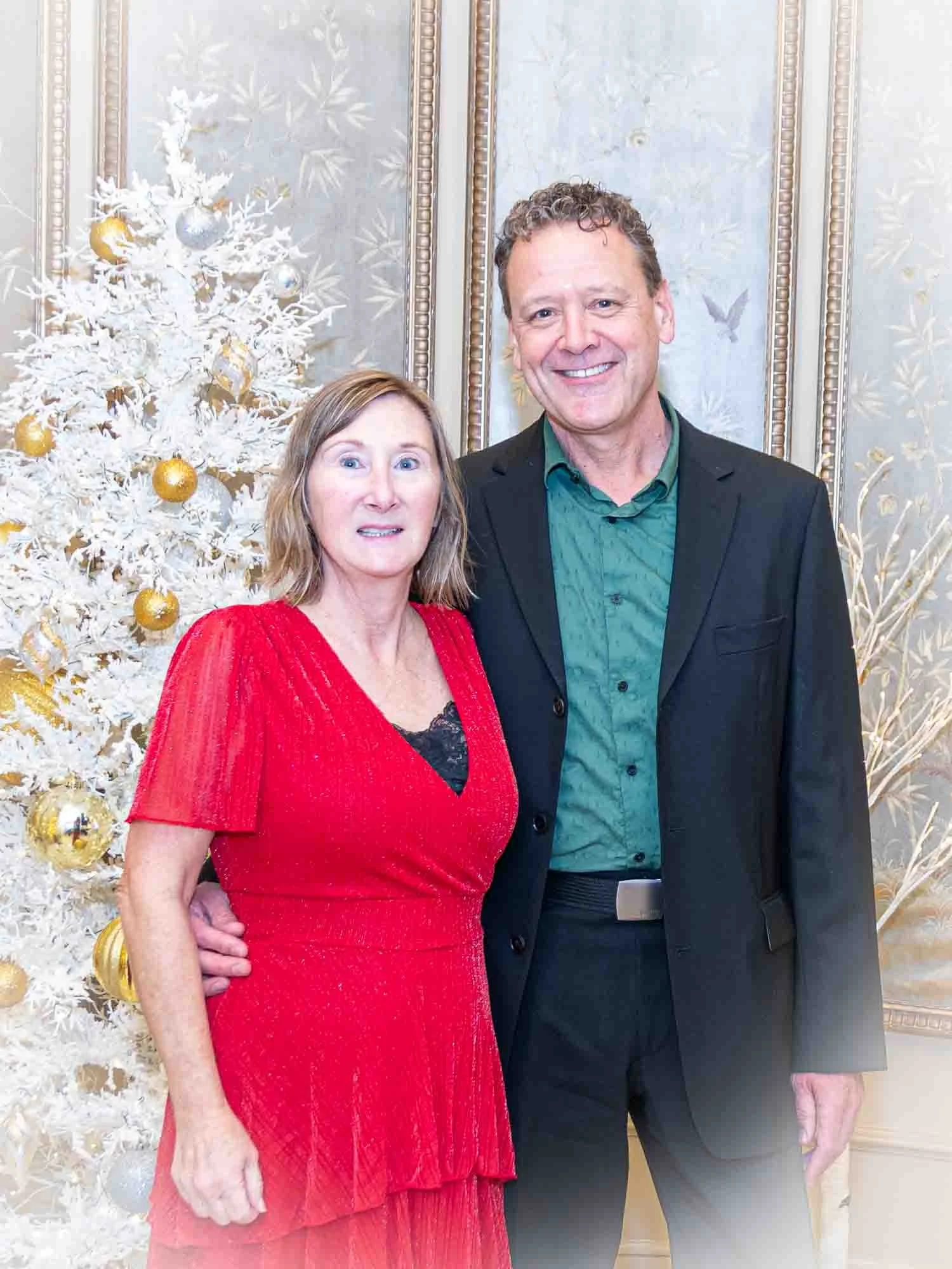 A smiling couple posing in front of a decorated white Christmas tree with gold and silver ornaments. The woman is wearing a red dress and the man is wearing a black suit with a green shirt.