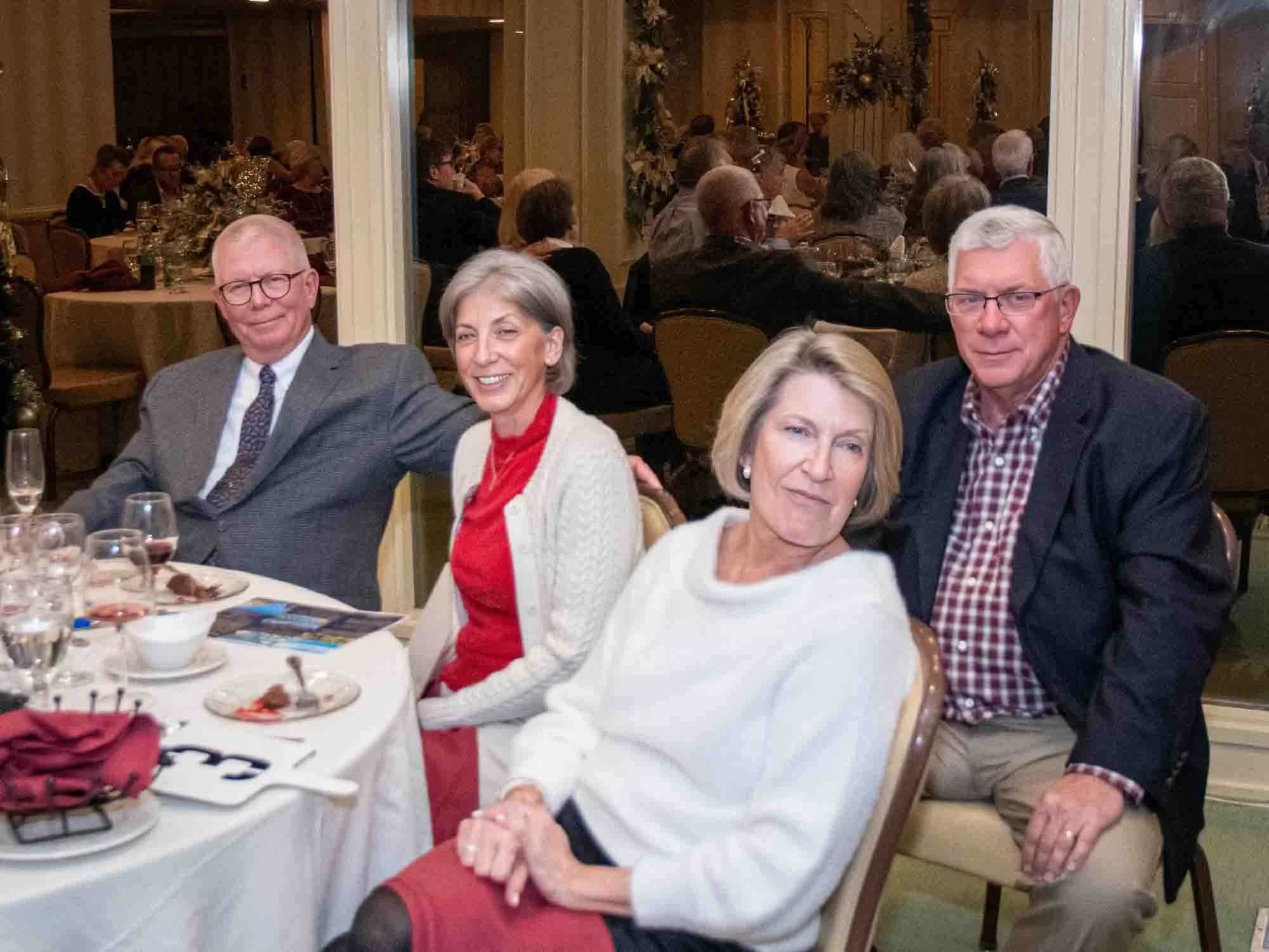Four people seated at a dining table in an evening event, with glasses, plates, and a napkin on the table.