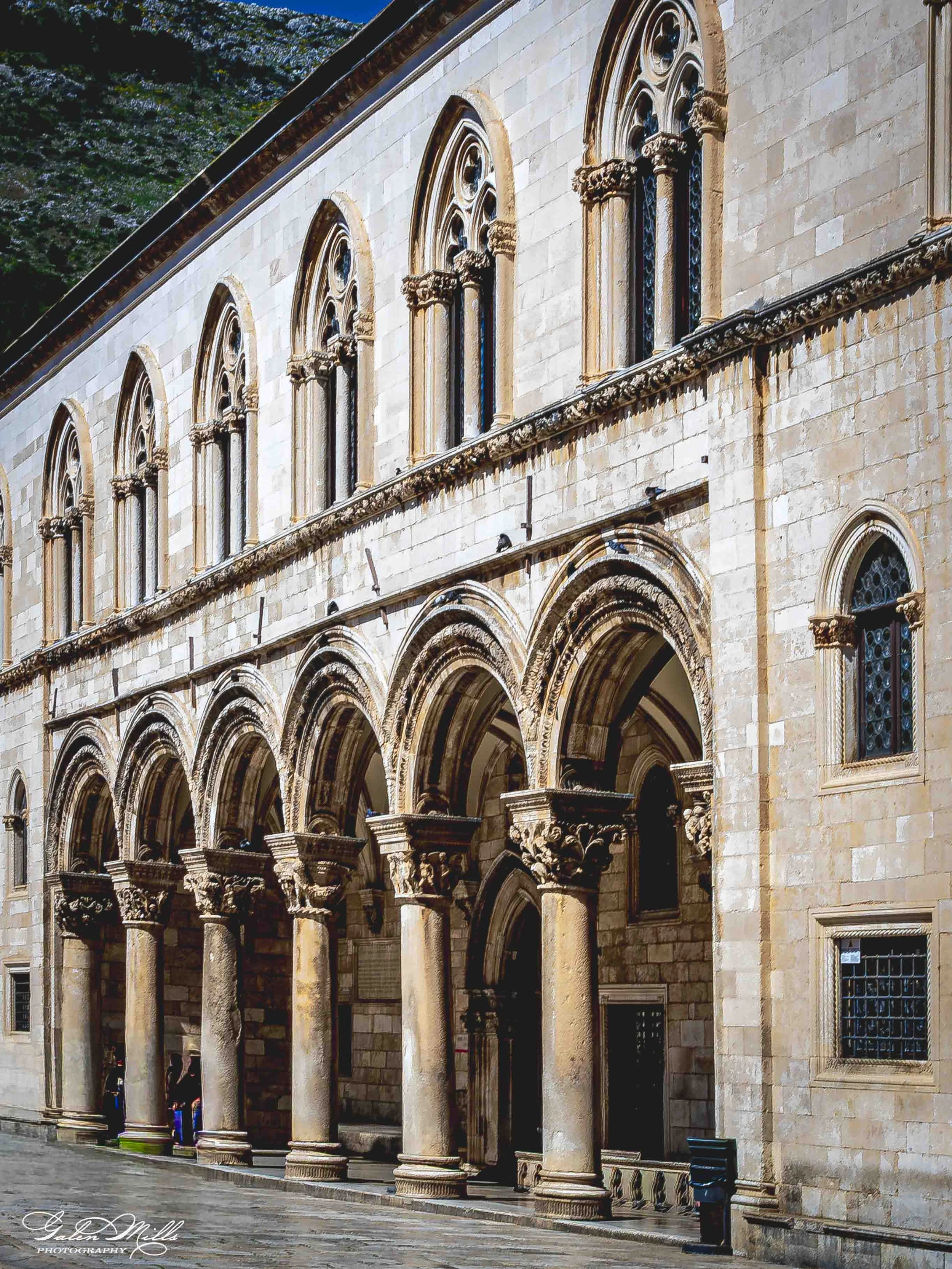 Exterior view of a historic stone building with arched doorways and tall narrow windows, featuring intricate stonework and columns, possibly a cathedral or church, with a paved street in front.