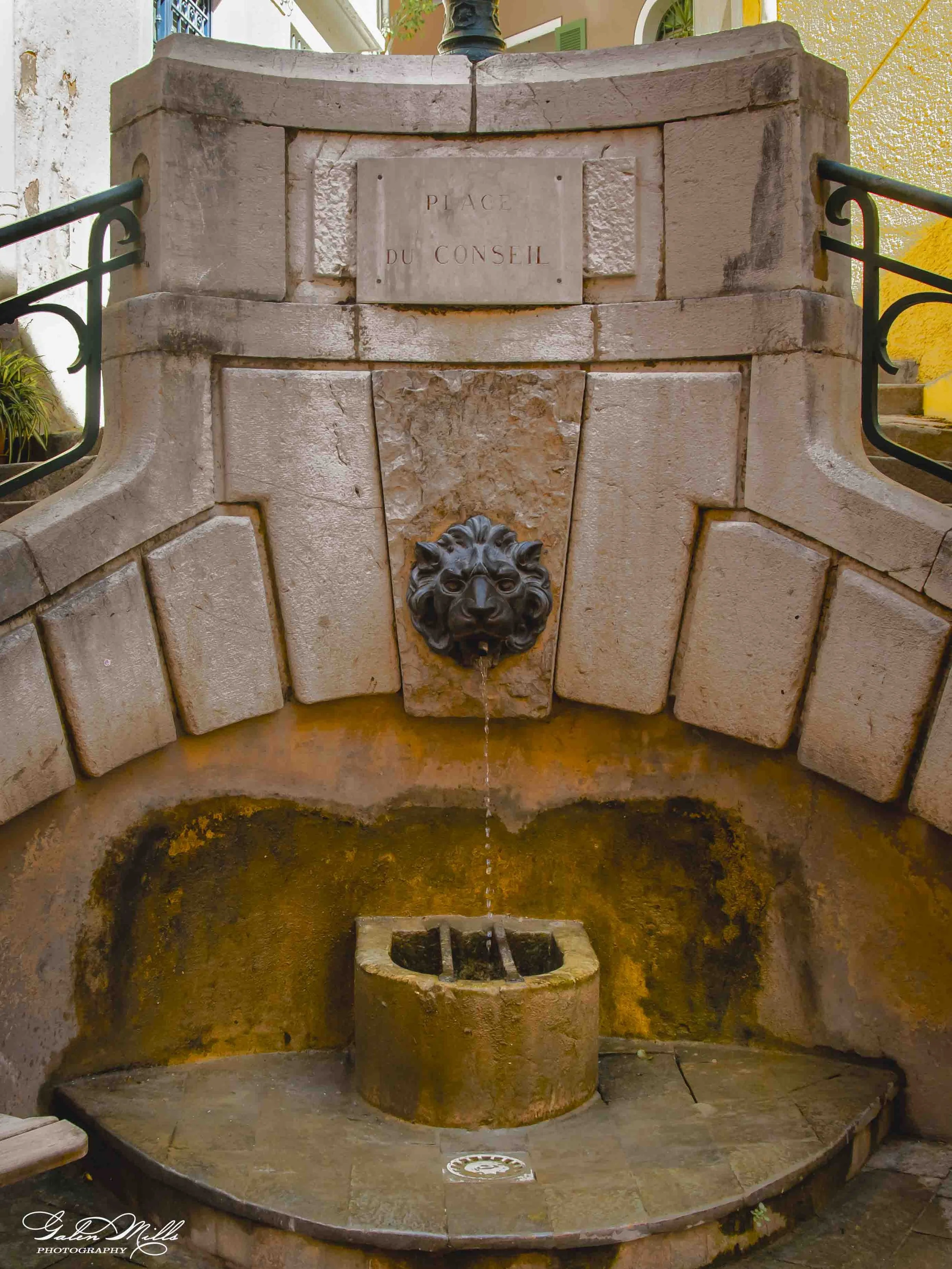 A stone fountain with a lion head water spout, a plaque that reads 'PLACE DU CONSEIL,' and a small basin at the top of the fountain. The fountain is situated outdoors with a wall and plants in the background.