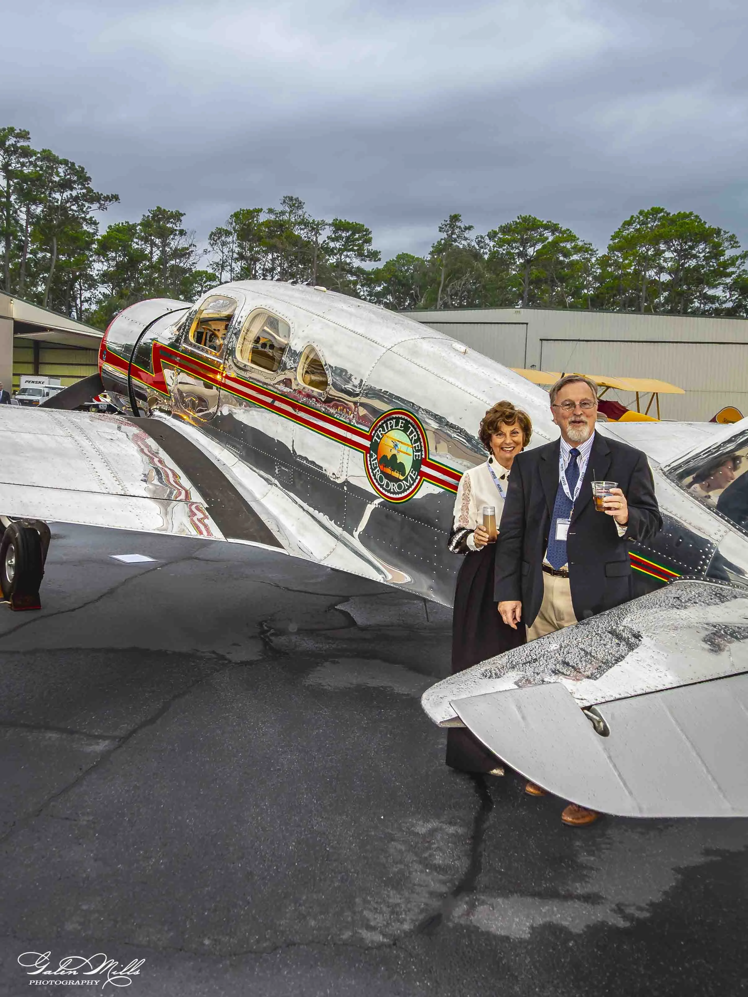 Two people standing in front of a shiny, vintage Silver Tri-Pacer airplane with a logo on its side, at an outdoor event under cloudy skies.