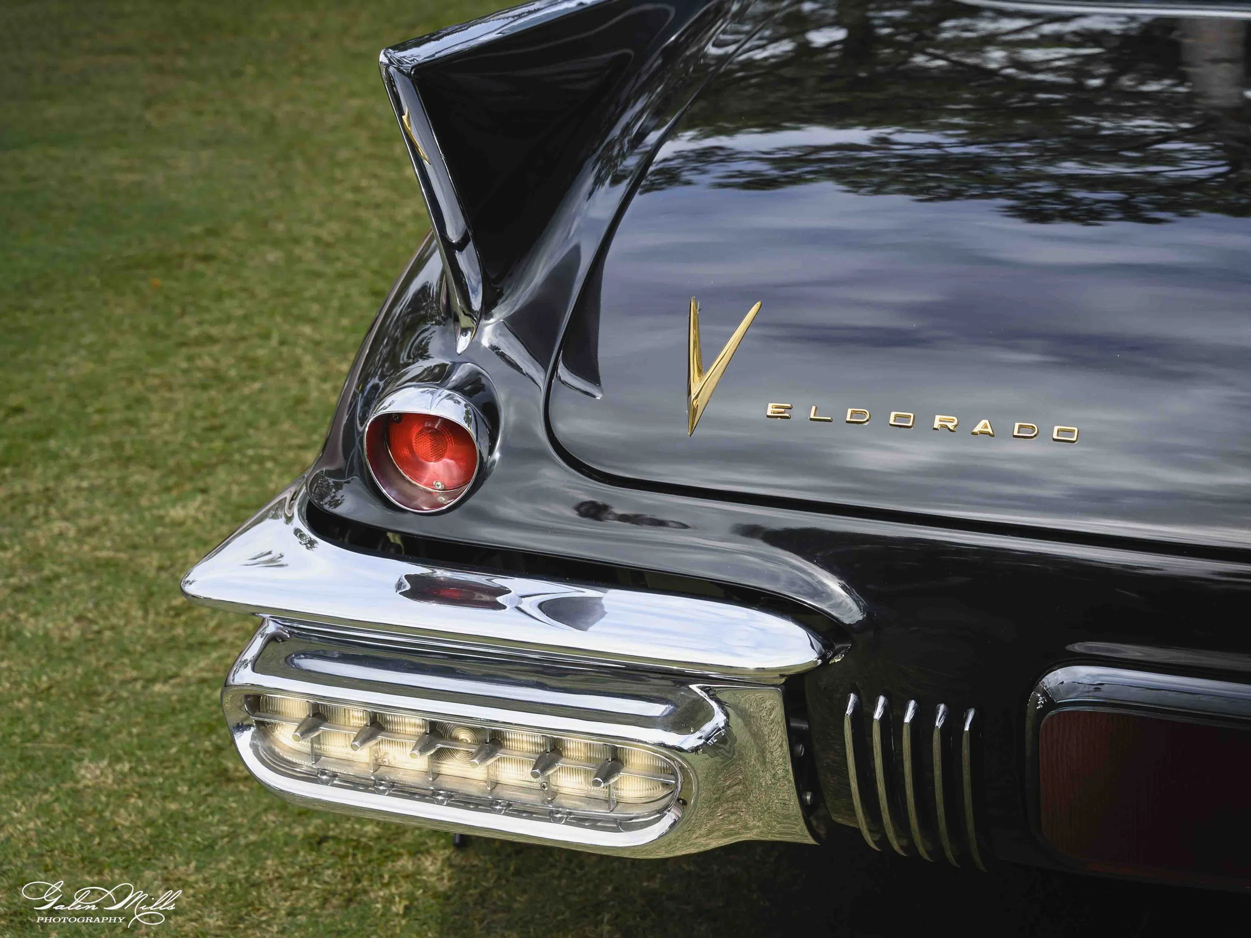 Close-up of the rear of a shiny black vintage Eldorado car, showing tail light, chrome accents, and the Eldorado emblem, parked on grass.