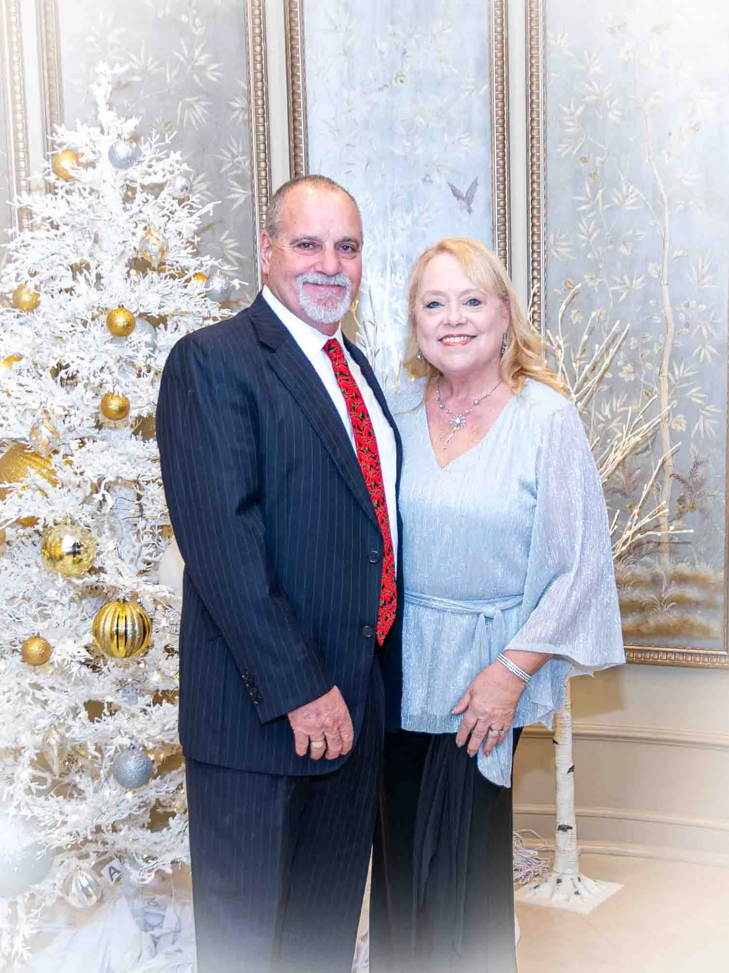 A man in a suit with a red tie and a woman in a light blue blouse, standing in front of a decorated white Christmas tree with gold and silver ornaments, in an elegantly decorated room.
