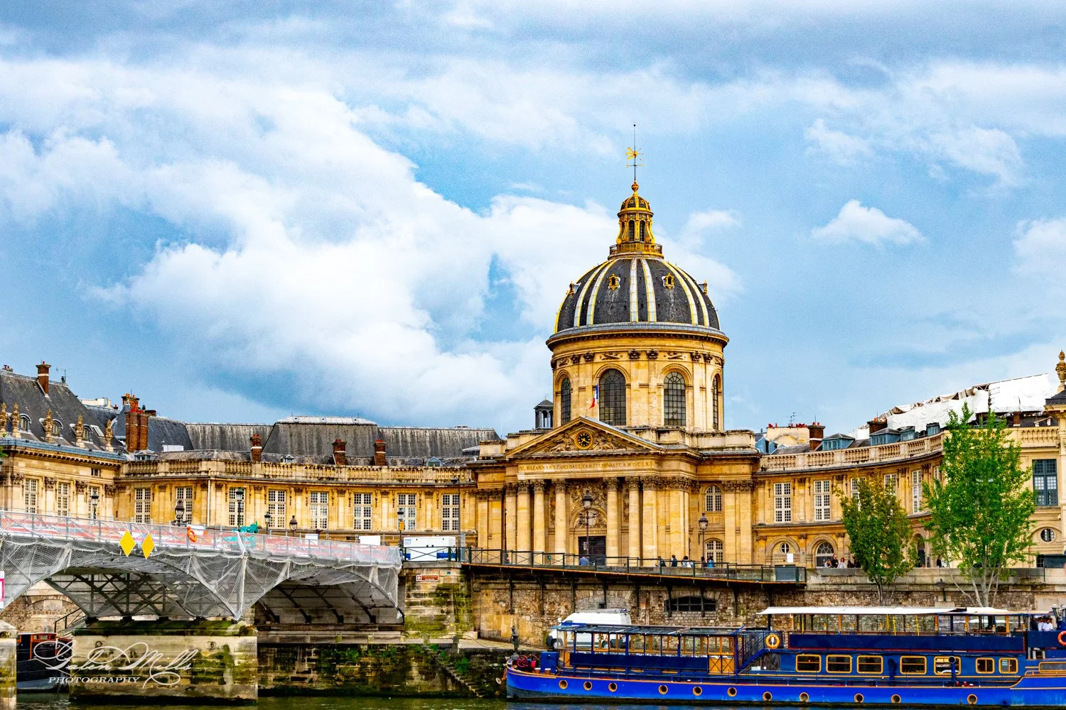 A large, historic domed building with classical architecture beside a river. In front of the building, there's a covered pedestrian bridge and a docked blue boat. The sky is partially cloudy.