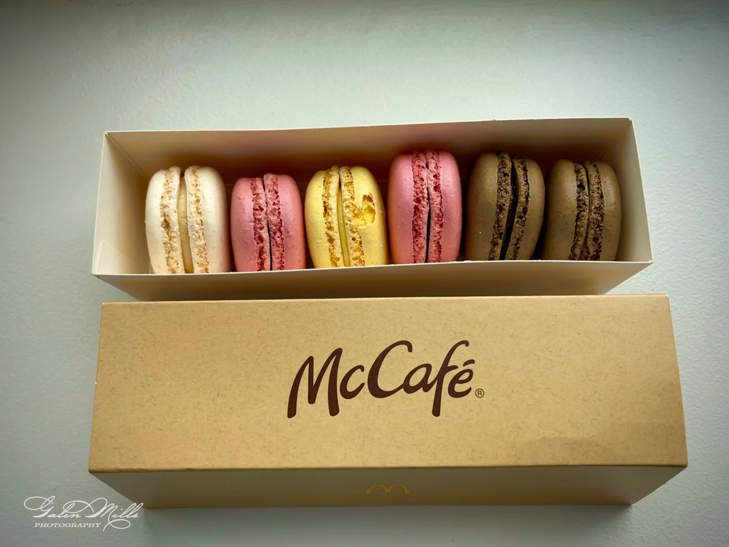 A box of assorted McCafé macarons, including white, pink, yellow, and brown flavors, displayed on a white surface.