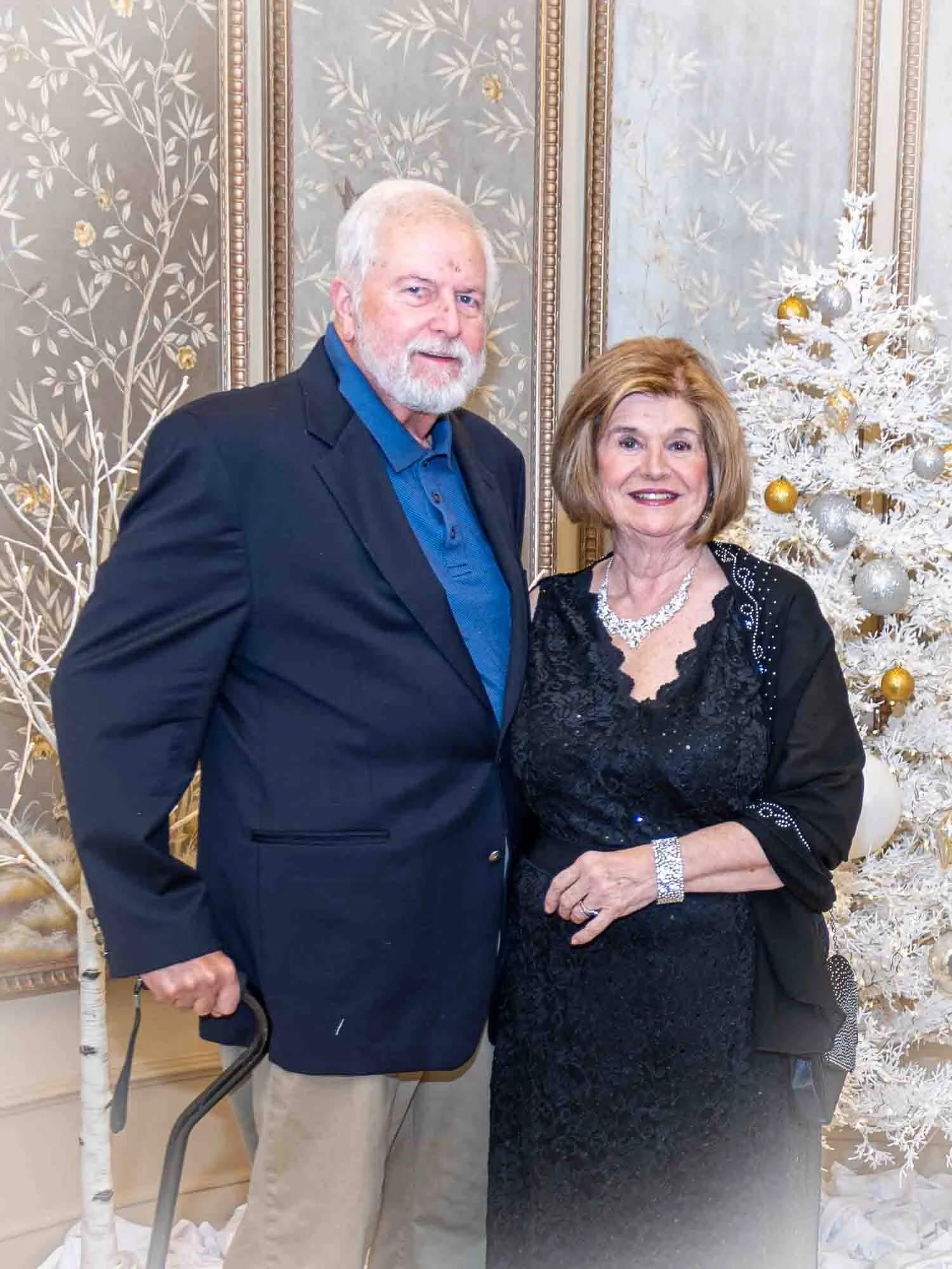 Elderly couple posing elegantly in formal attire, standing next to a decorative white Christmas tree adorned with gold and silver ornaments, in front of an ornate, patterned wall.