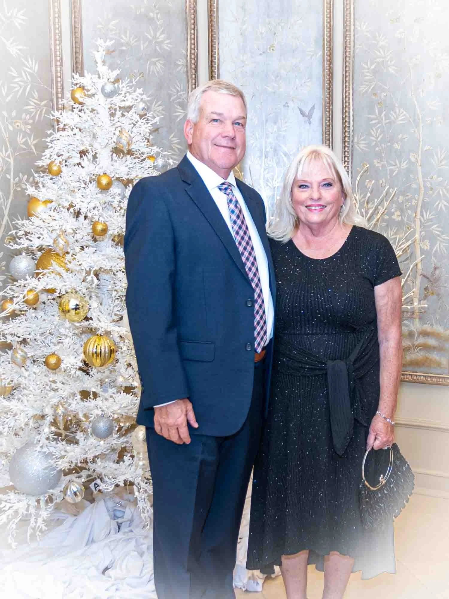 Couple posing in front of a decorated white Christmas tree with gold and silver ornaments in an elegantly decorated room; man in a suit and woman in a black dress.