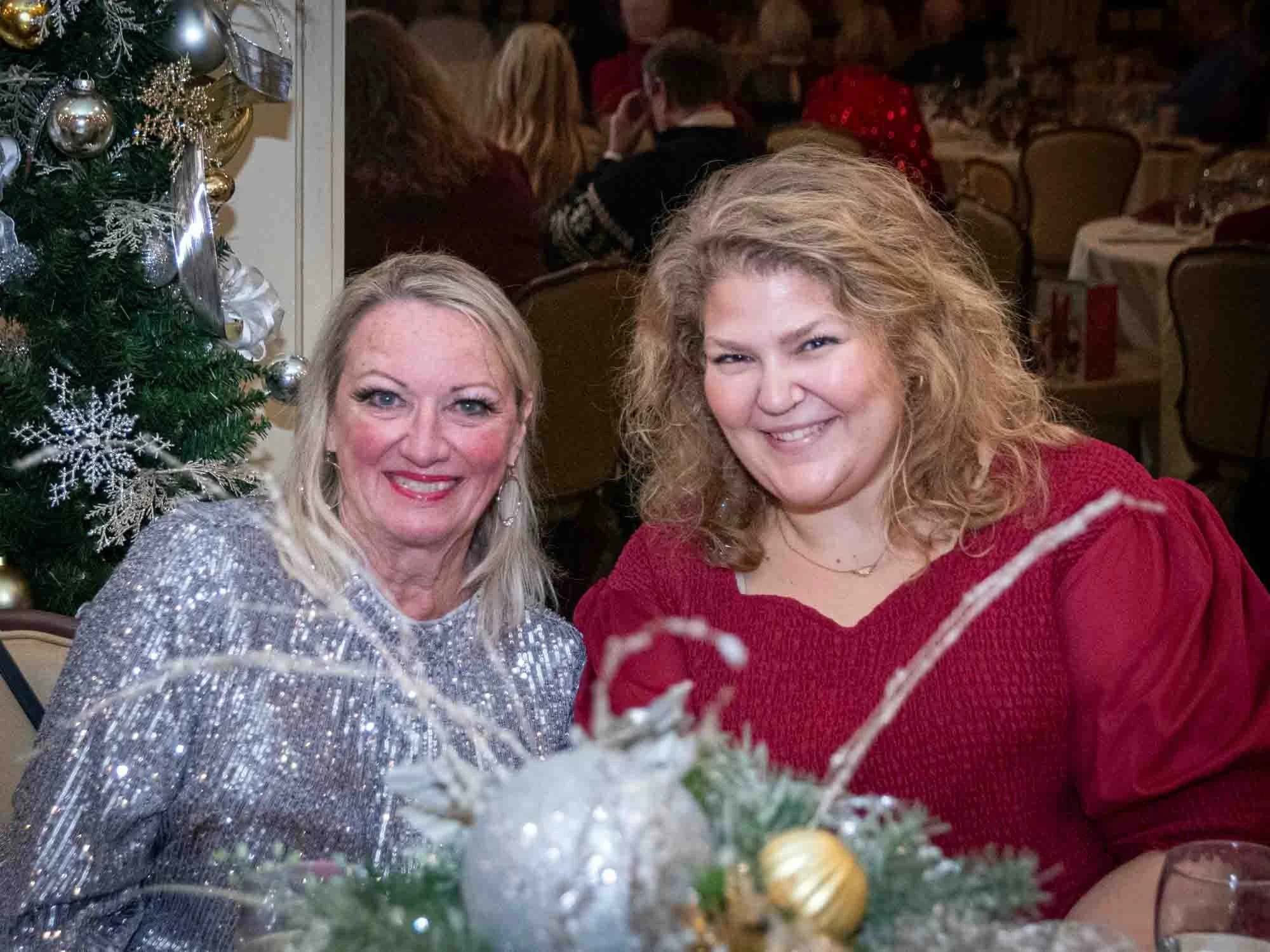 Two women smiling at a festive event, with a Christmas tree and decorations in the background.