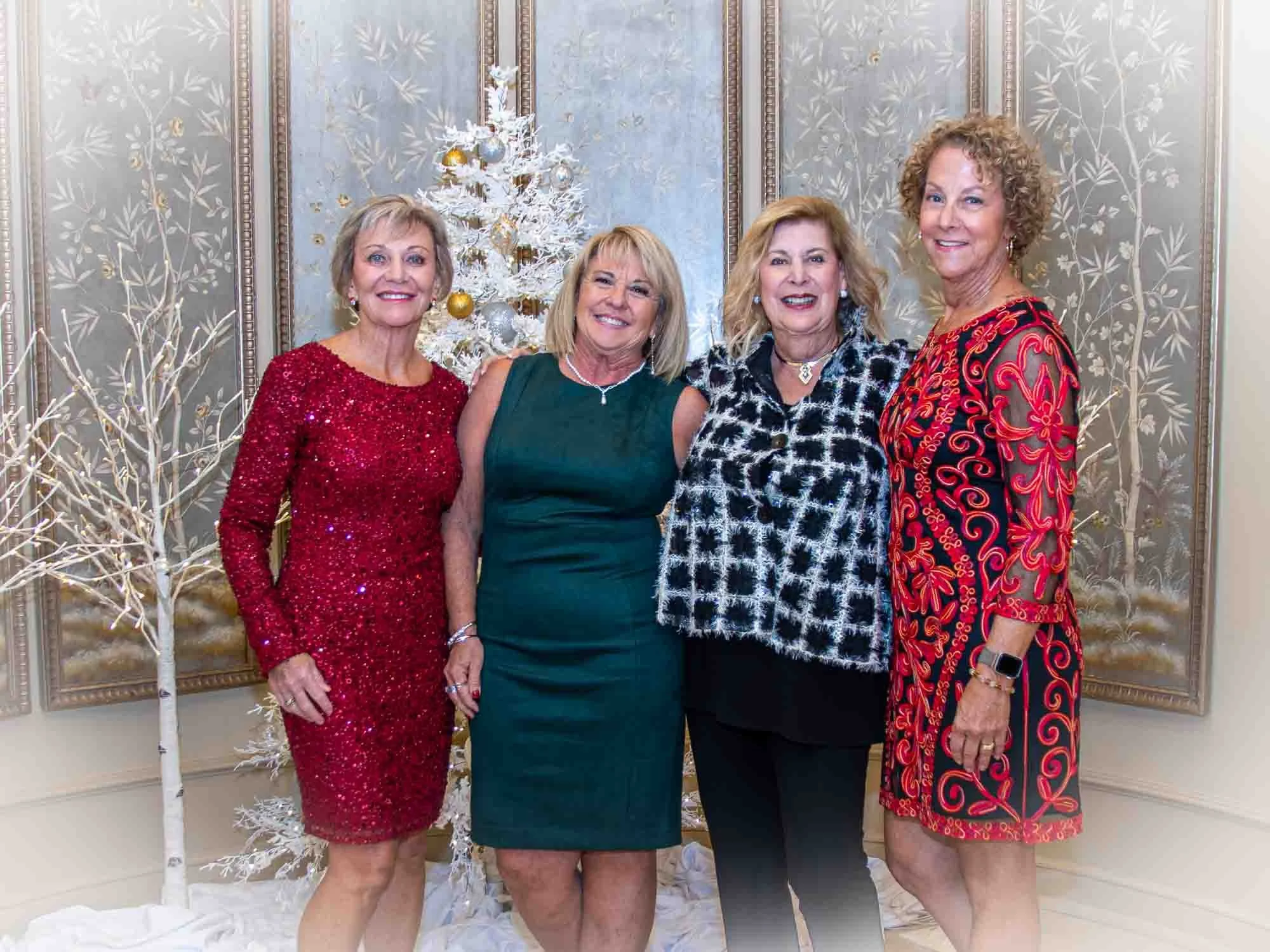 Four women posing together at a festive event with a decorated white Christmas tree and elegant backdrop.