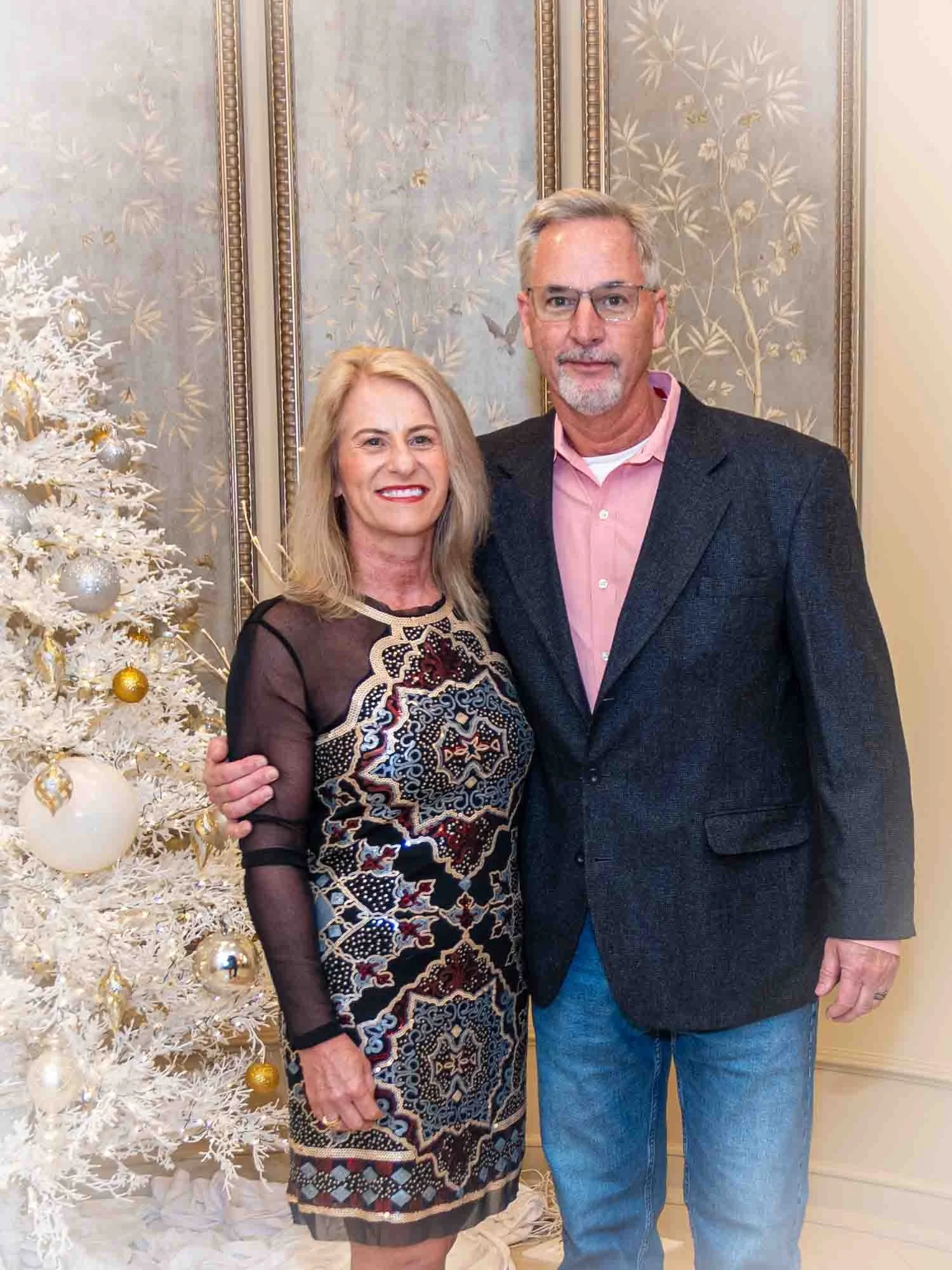 A couple posing indoors near a decorated white Christmas tree. The woman is wearing a patterned dress and the man is wearing a dark blazer with jeans. The background features ornate paneled walls.