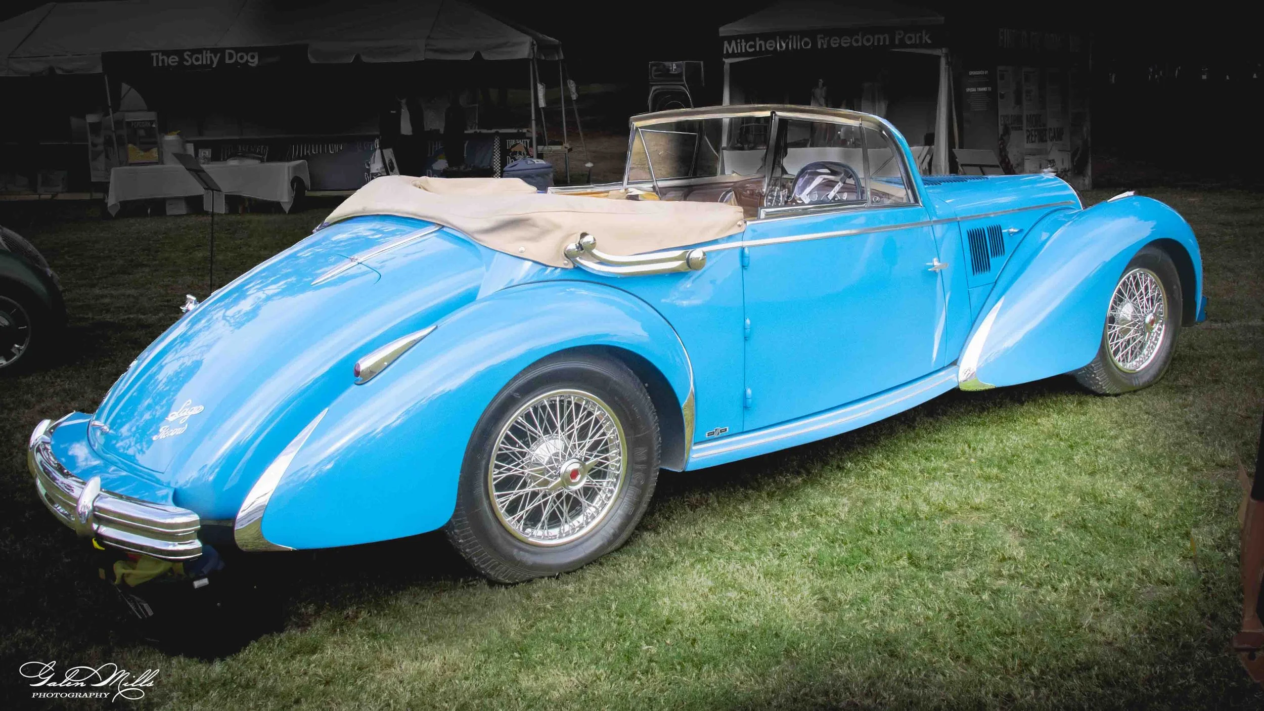 A vintage blue convertible car with beige interior and wire wheels, parked on grass at an outdoor event with tents and booths in the background.