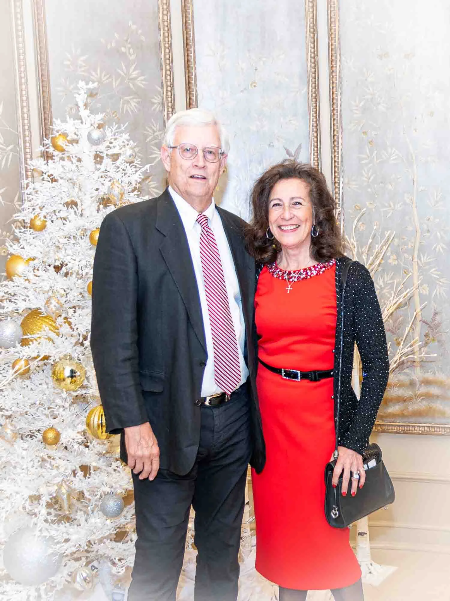 A man and a woman posing in front of a decorated white Christmas tree with gold and silver ornaments. The man is wearing a dark suit with a striped tie, and the woman is in a red dress with a black belt and a black purse.