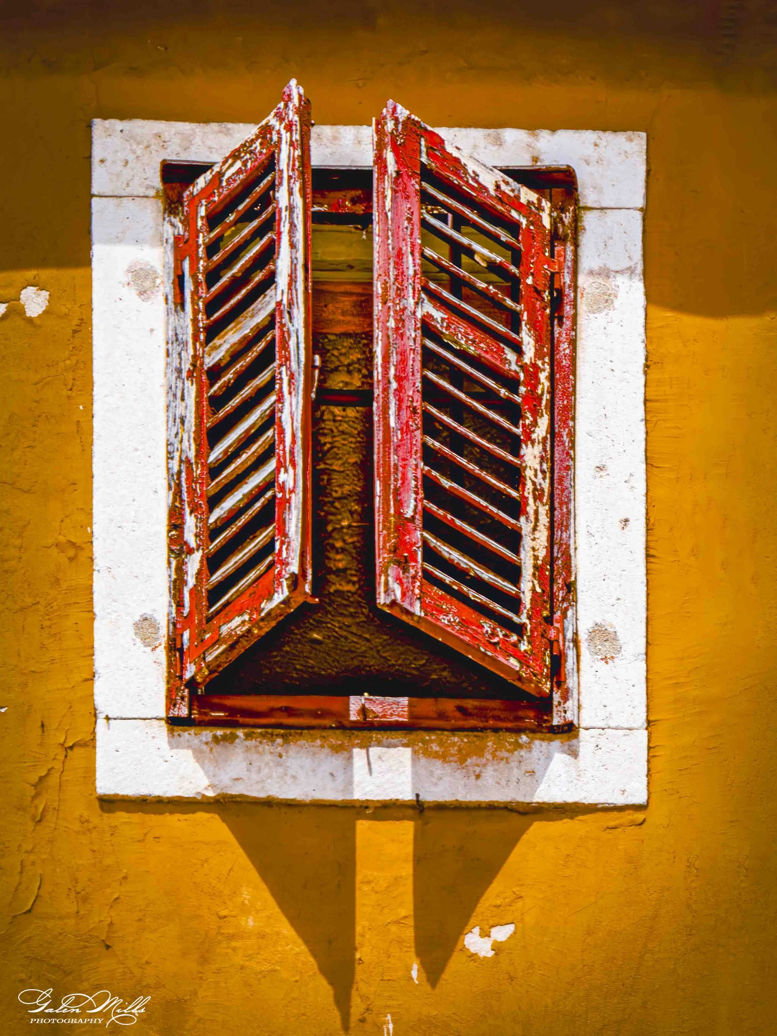 Weathered red wooden window shutters mounted on a yellow stucco wall, with painted white trim around the window, casting a shadow downward.