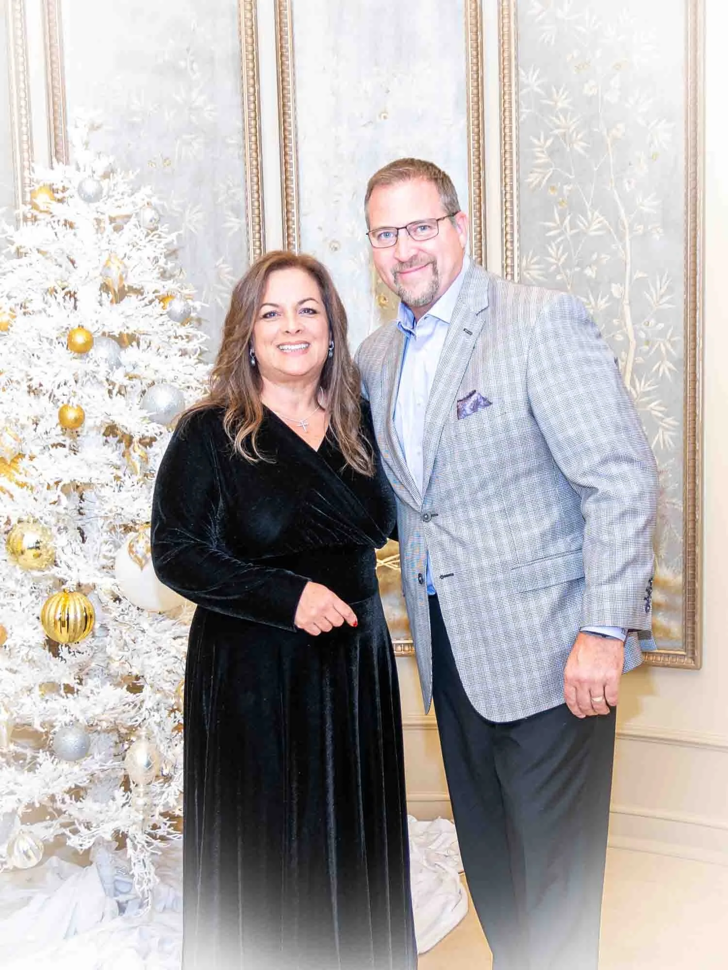 A man and woman standing together in front of a white Christmas tree with gold and silver ornaments, inside a decorated room.