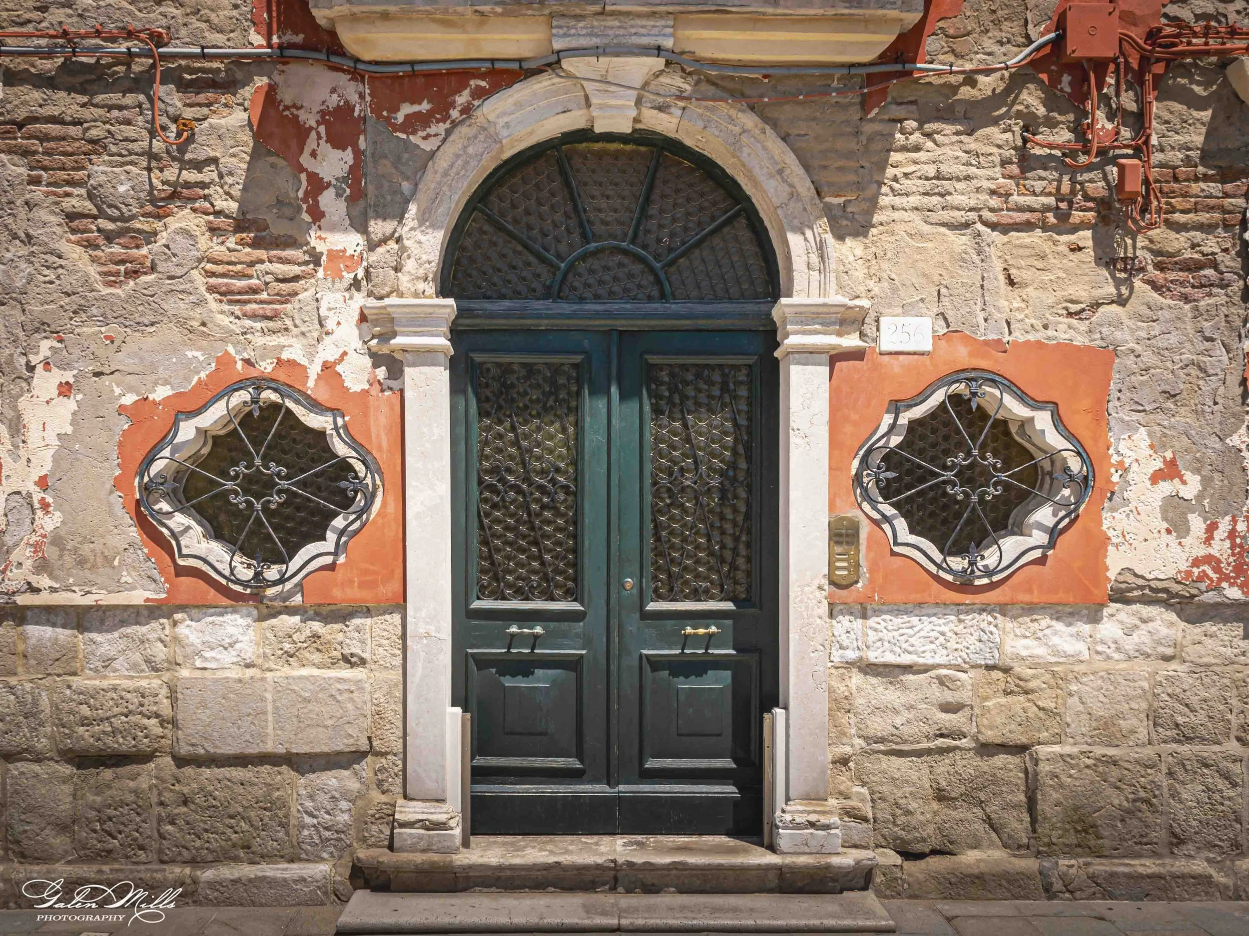 A black double door with decorative ironwork is set in an aging stone and brick wall with patches of peeling paint. Flanking the door are two small oval windows with ornate wrought iron grilles, and a small rectangular house number plaque reads '256'