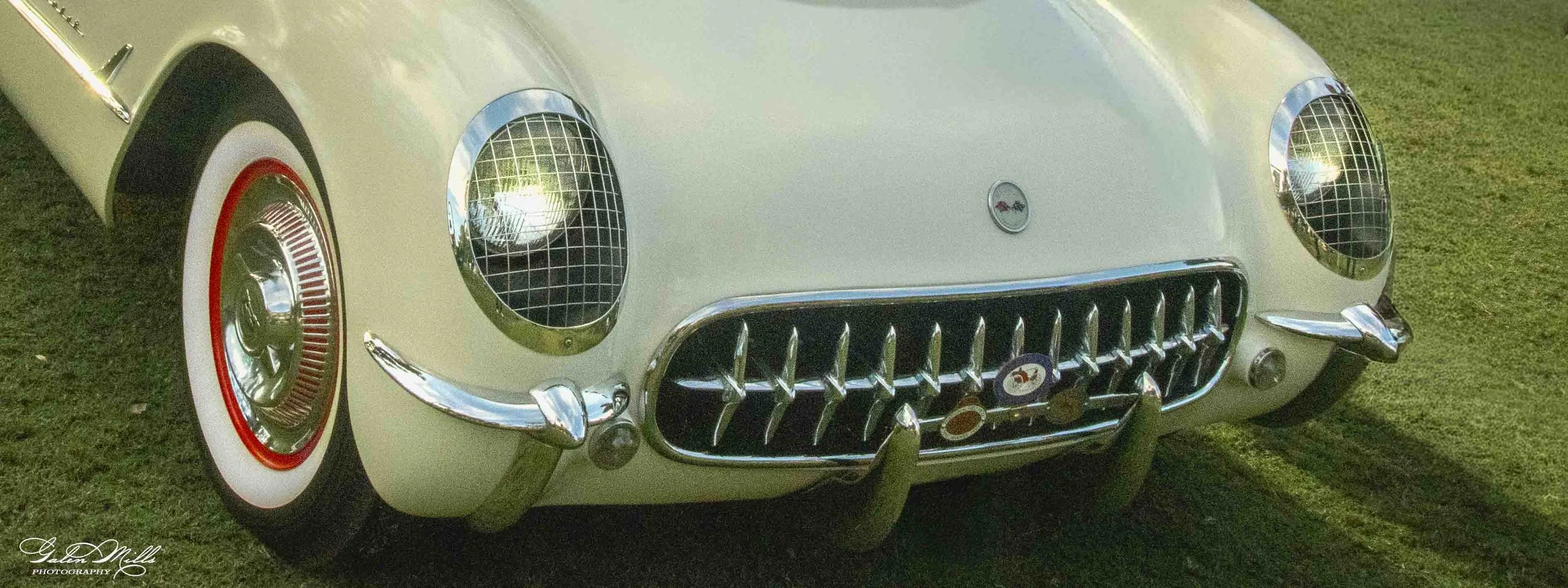Close-up of a vintage cream-colored car with chrome accents, showing the front grille, headlights with wire covers, and a red, white, and black hubcap on the front wheel, parked on grass.