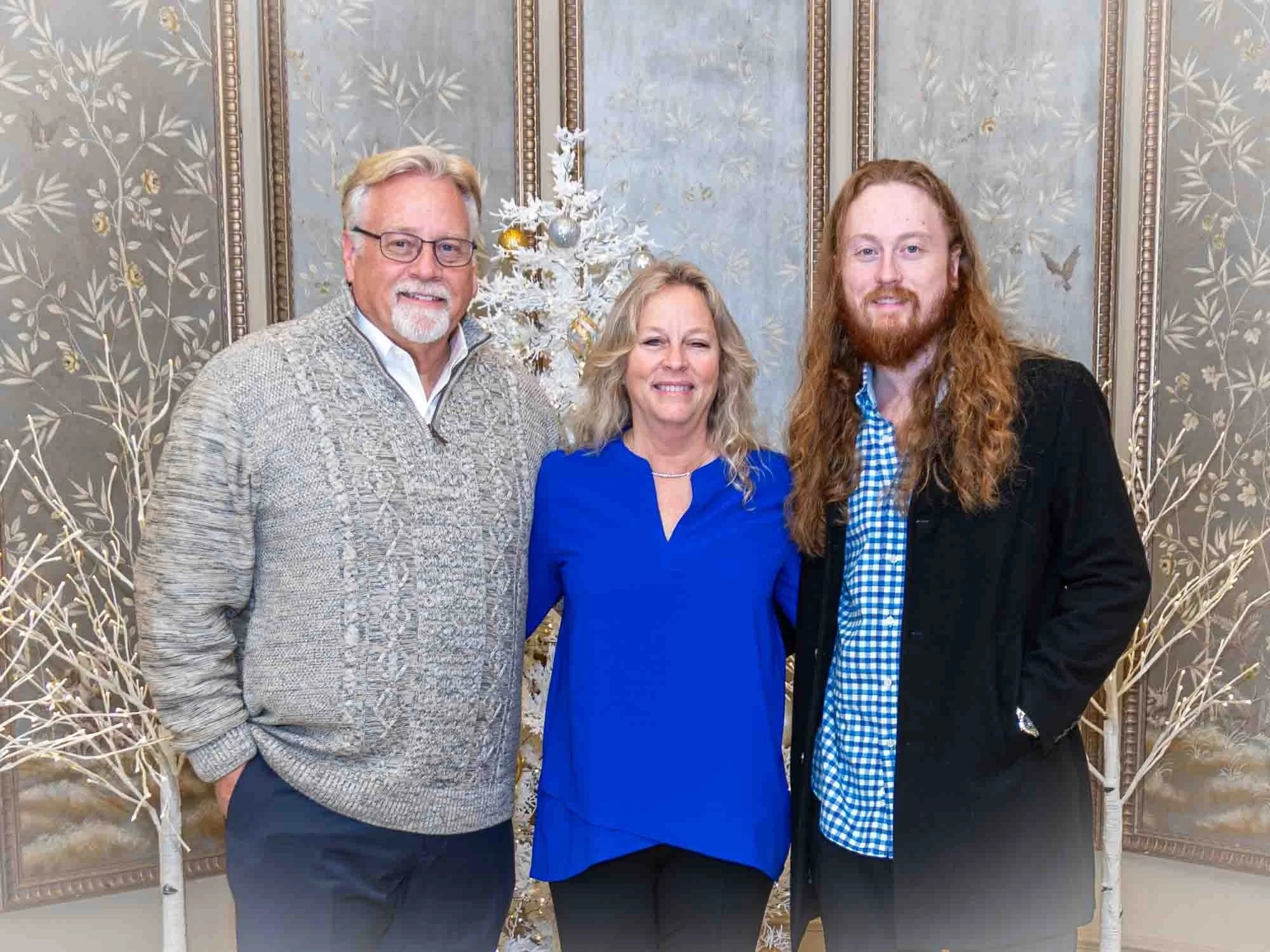 Three people posing in front of a decorative backdrop with white trees, one man wearing a gray sweater, a woman in a blue blouse, and another man in a black jacket.