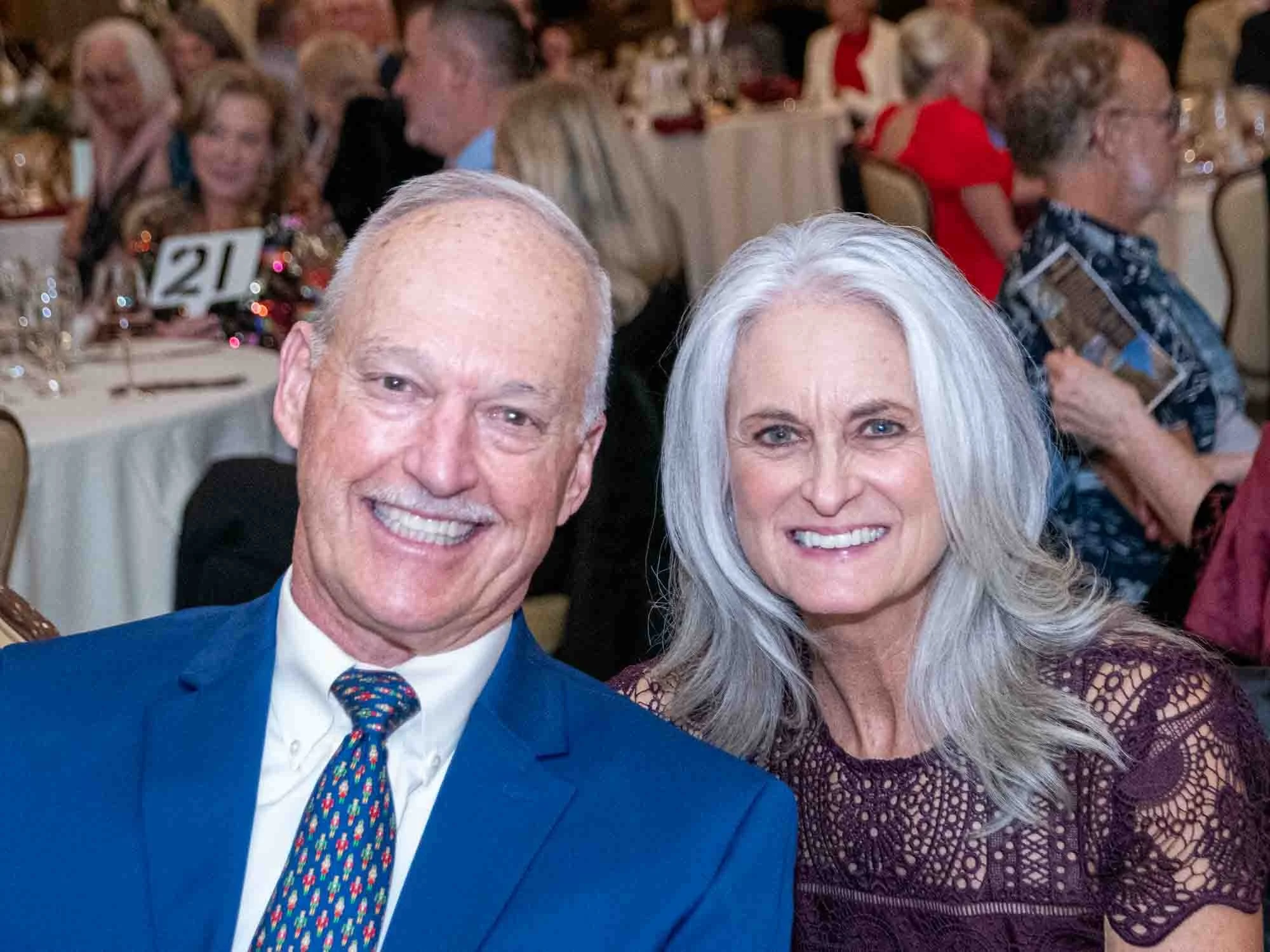 A smiling man in a blue suit and patterned tie sits next to a smiling woman with long gray hair in a lace dress. They are at an event with tables and attendees visible in the background.