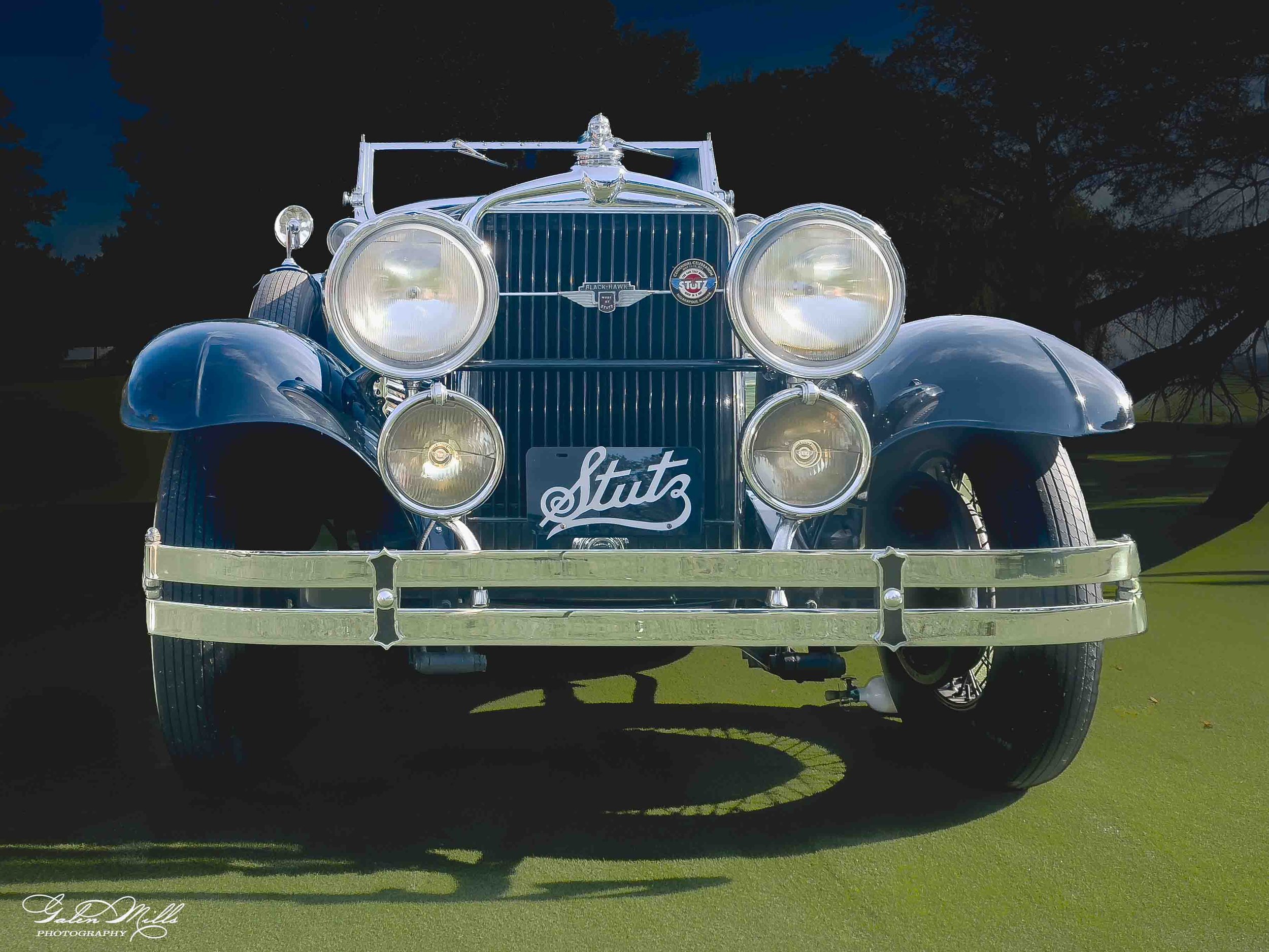 Front view of a vintage silver car parked on grass with a dark blue sky and trees in the background.