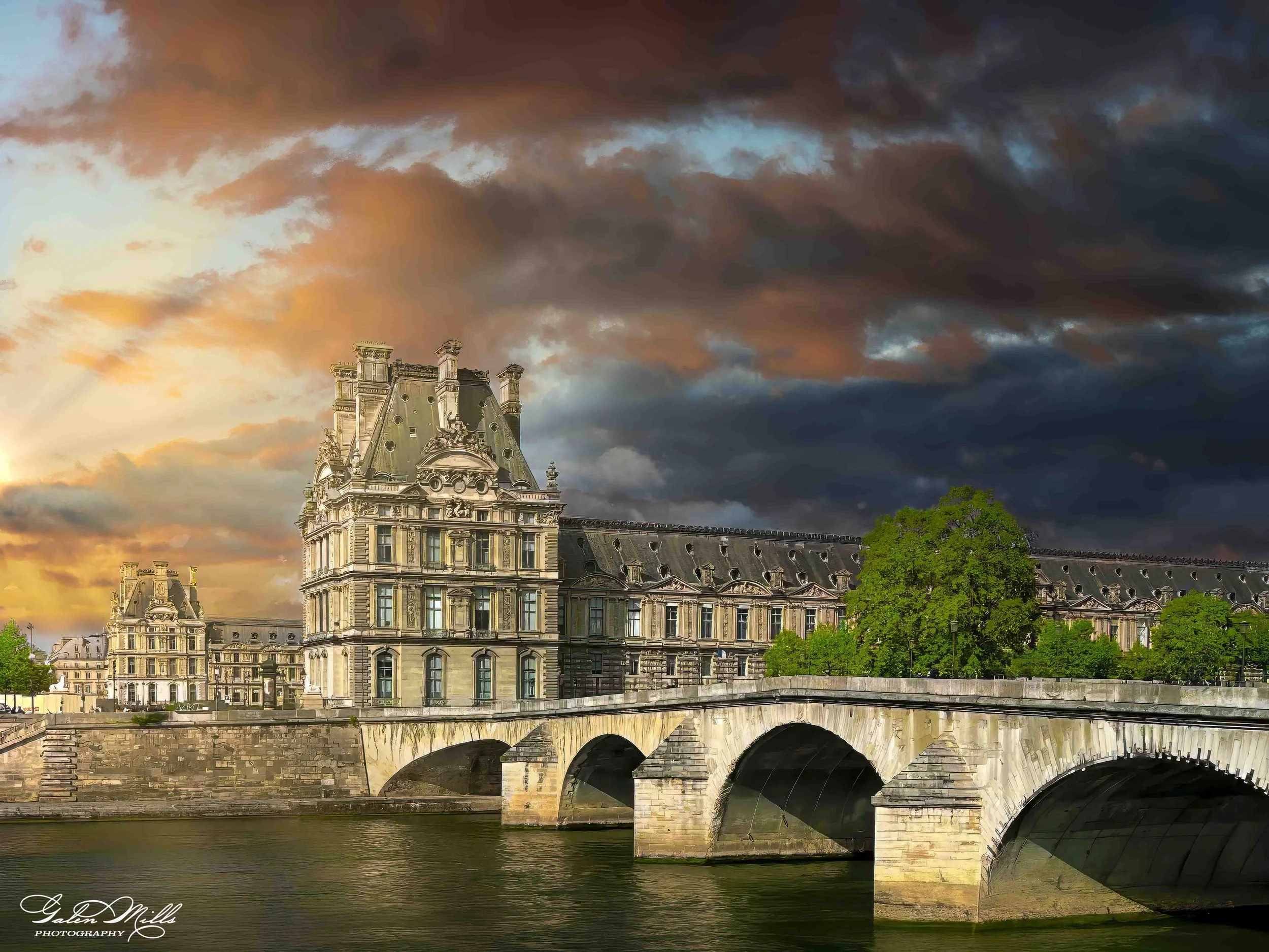 Historic building and bridge over tranquil river at sunset with dramatic clouds.