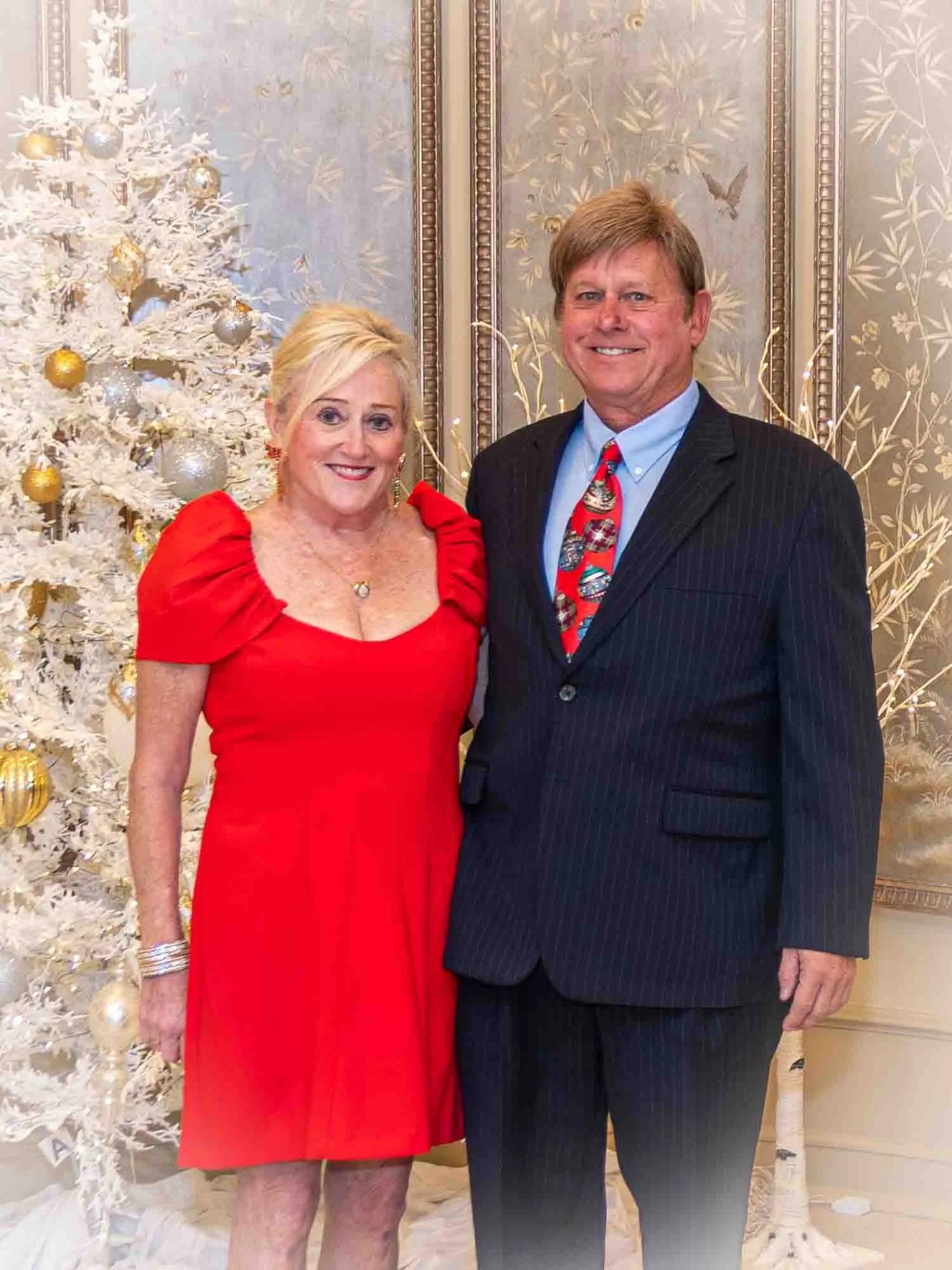 A woman in a red dress and a man in a suit with a festive tie standing in front of a decorated white Christmas tree.