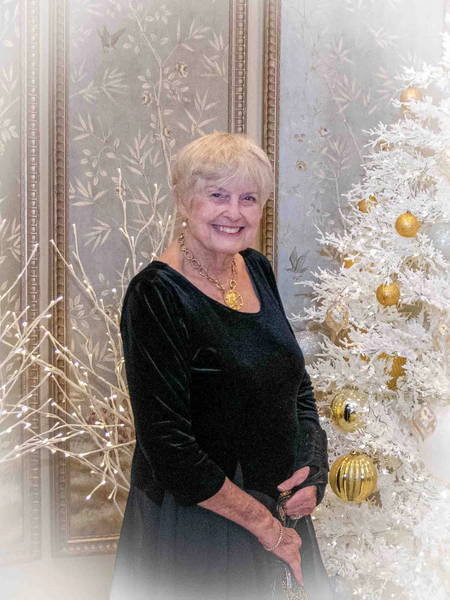 Elderly woman in a black dress standing beside a white Christmas tree with gold ornaments.