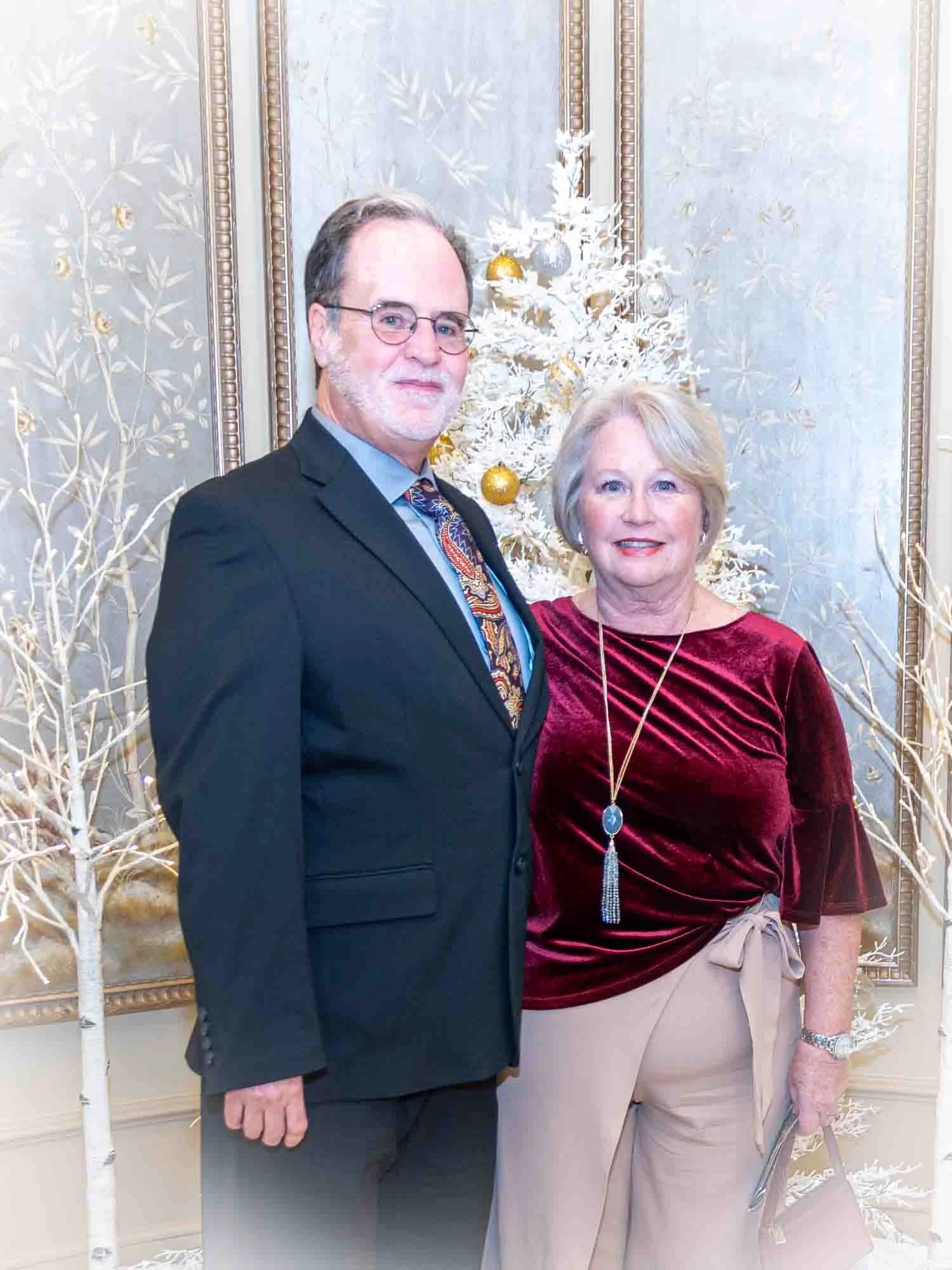 A couple posing in formal attire; the man wears a dark suit with a patterned tie and glasses, while the woman wears a burgundy blouse and beige pants. A white decorative tree stands in the background.