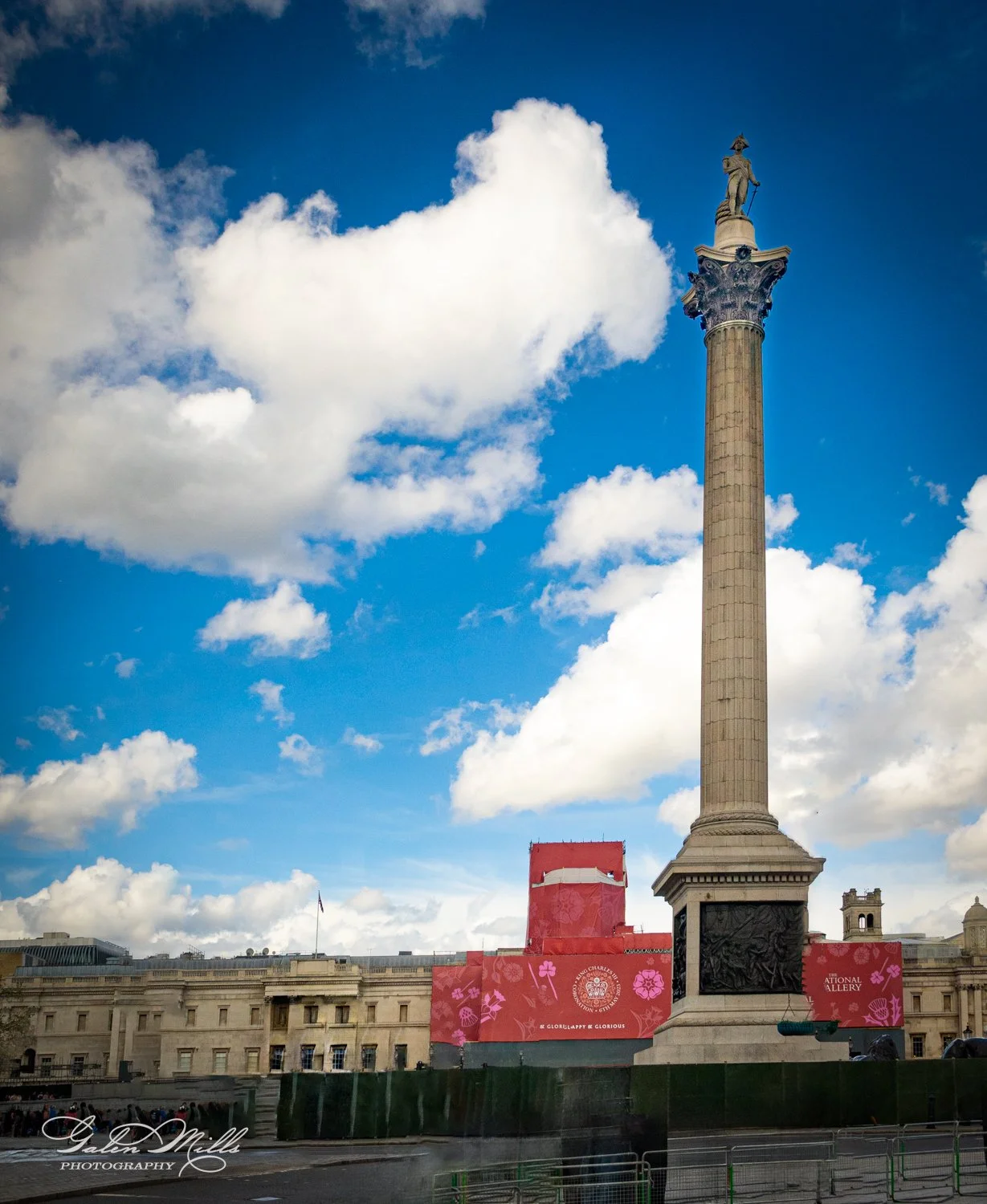 Tall column with a statue on top, surrounded by historical architecture and decorative banners, under a blue sky with clouds.