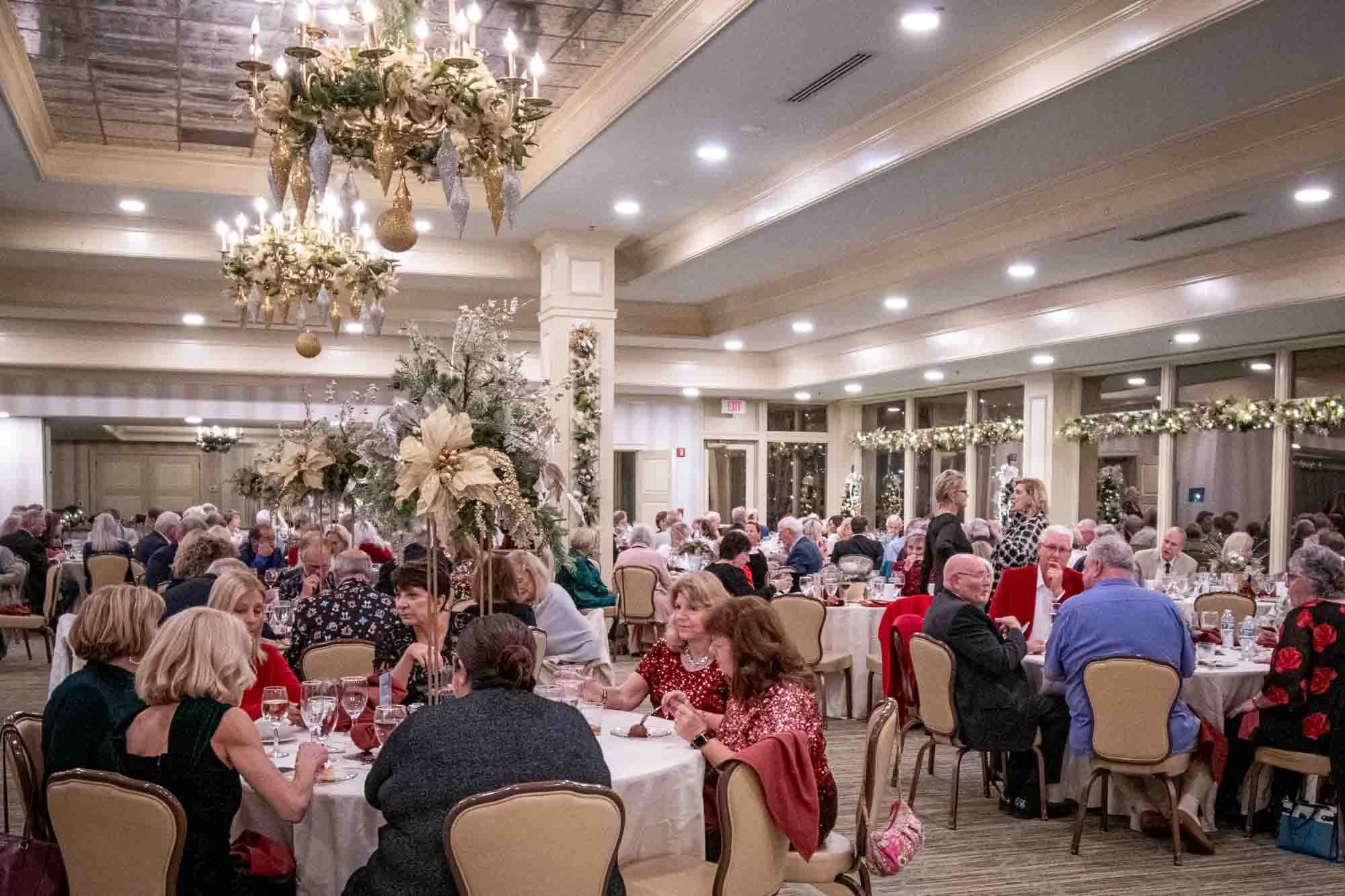 A large indoor gathering with people seated around tables under decorated chandeliers, likely a formal event or celebration. The room is adorned with festive decorations, including floral centerpieces and garland.