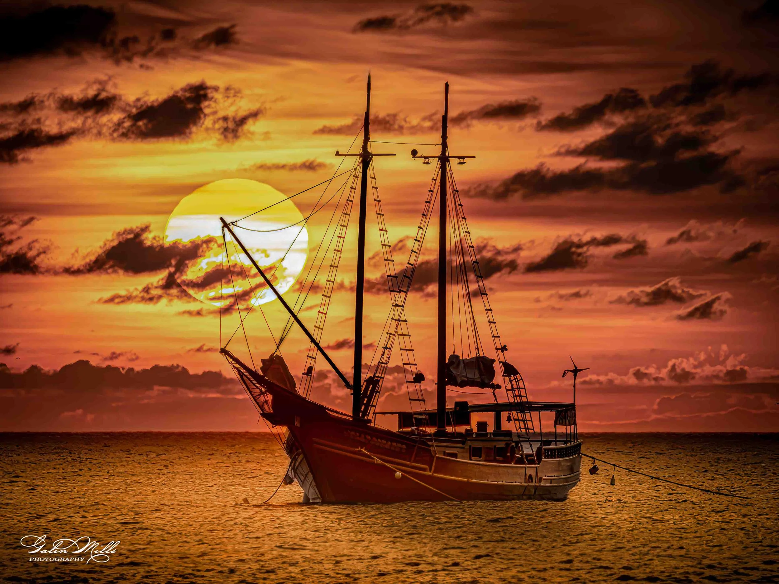 'A sailboat anchored on the water during a sunset with the sun low on the horizon and a sky filled with clouds in shades of orange, yellow, and dark brown.