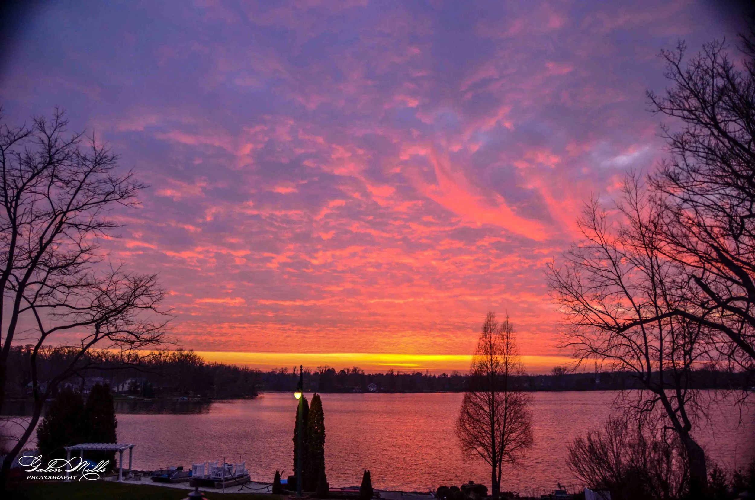 Sunset over a lake with colorful pink, purple, and orange clouds, silhouetted trees, and boats docked along the shore.