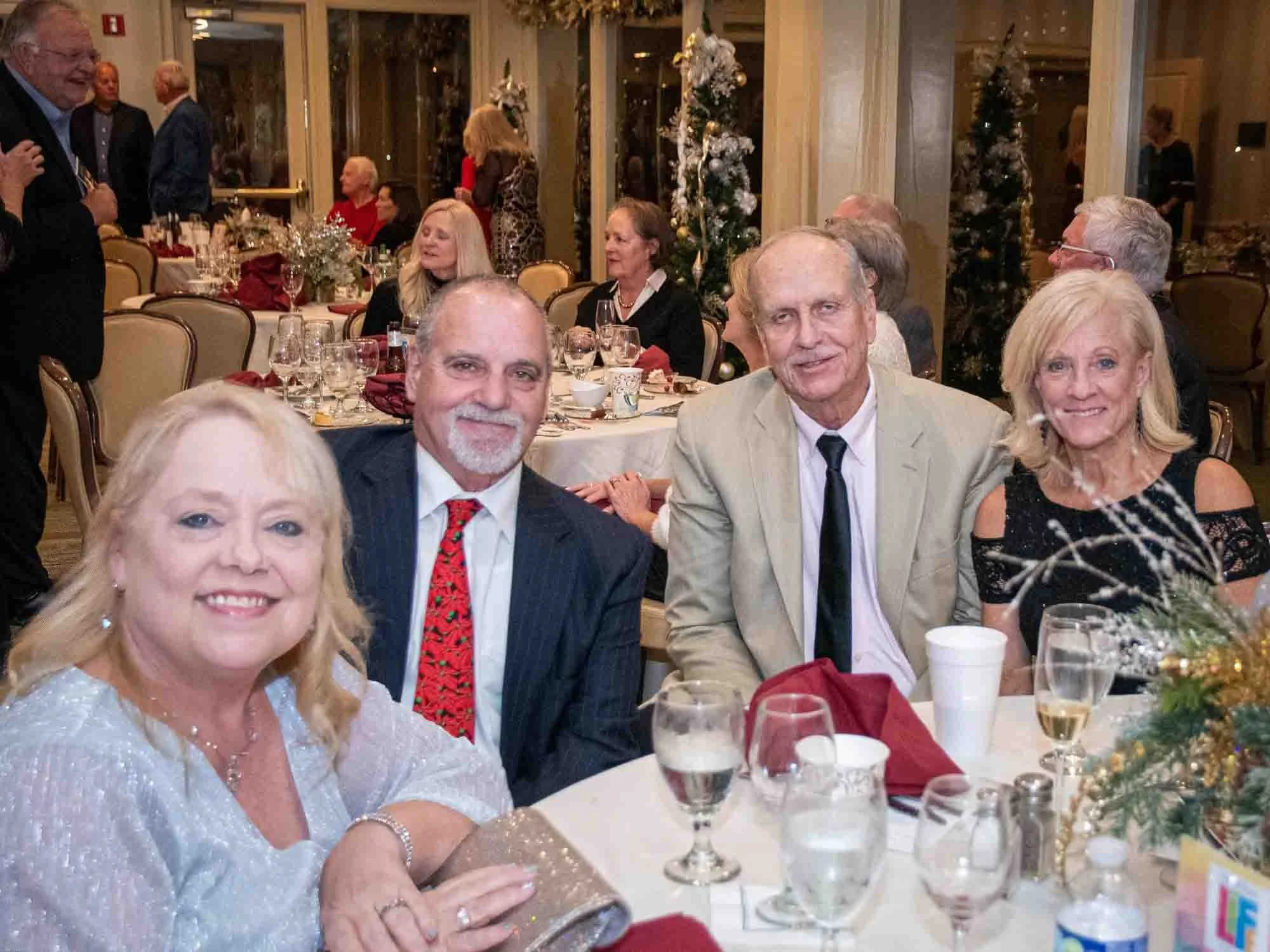 A group of people at a formal event, sitting at a table with festive decorations and drinks. Others are seated and mingling in the background.