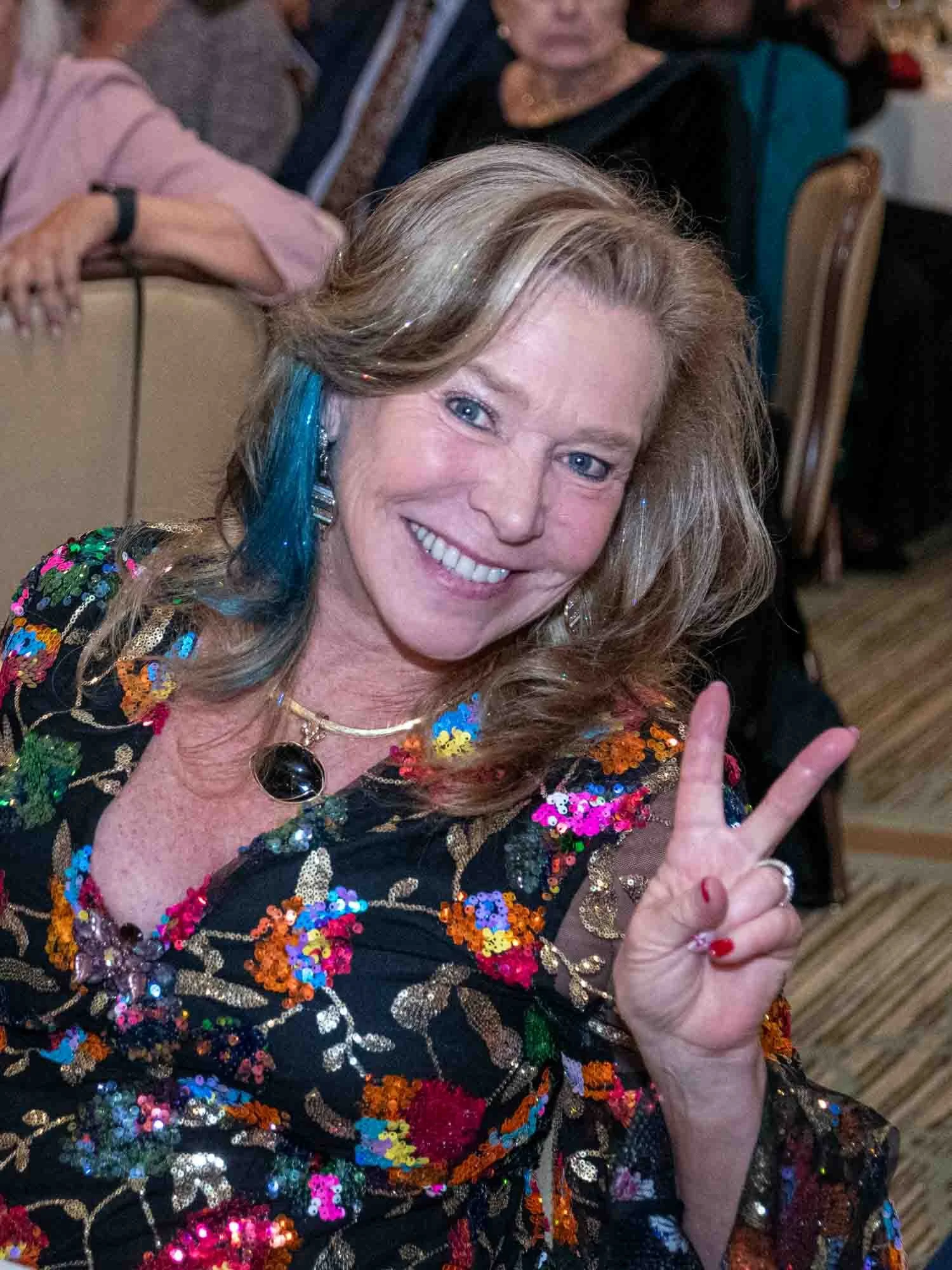 Smiling woman flashing a peace sign at an event, wearing a colorful sequined dress.