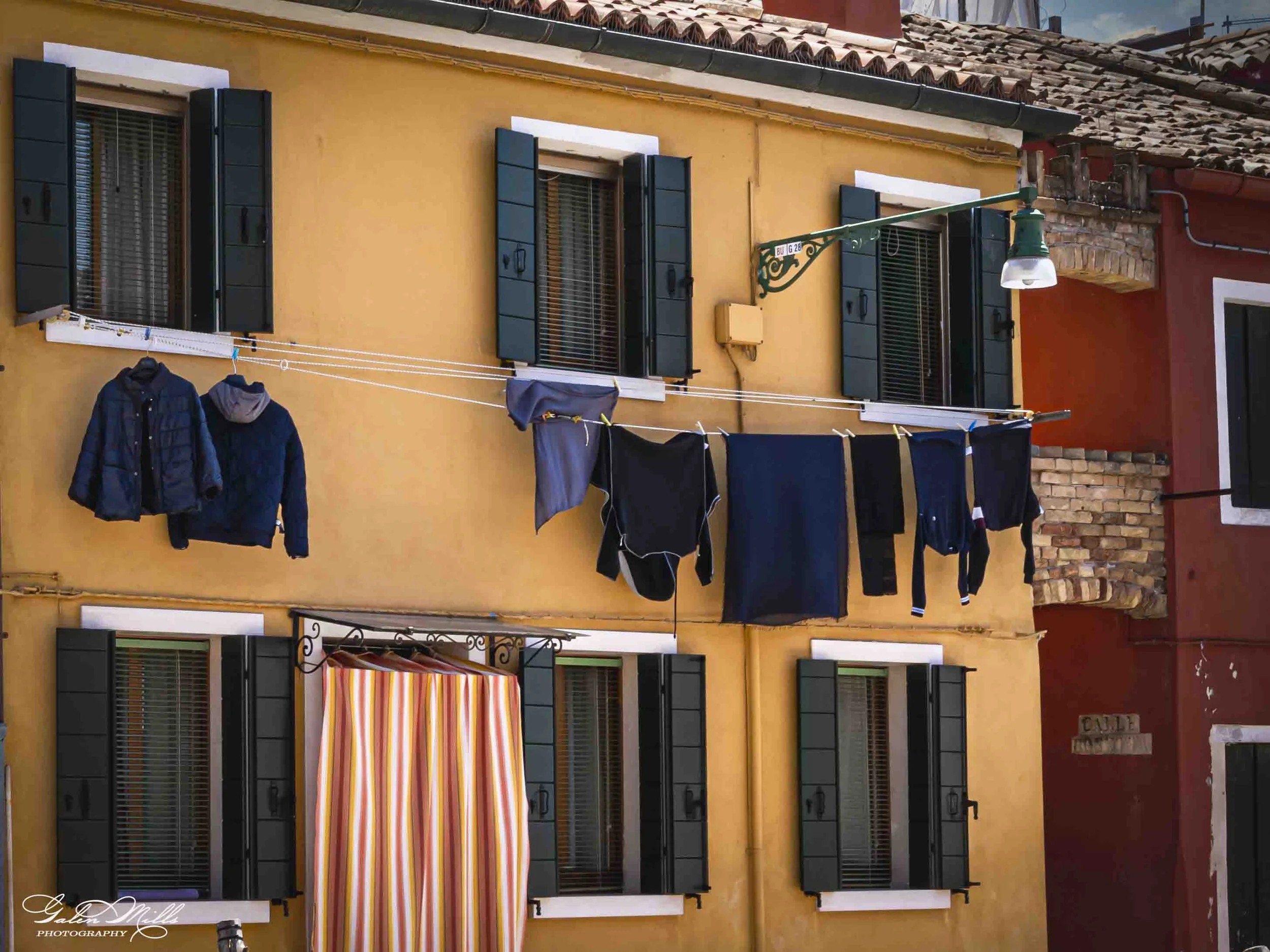 The image shows a yellow building with four windows that have black shutters and brown window frames. There are laundry lines with clothes hanging outside, including jackets, pants, and shirts. An ornate green street lamp is attached to the wall, and