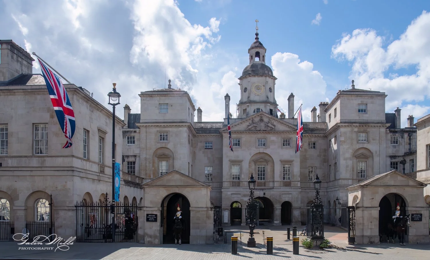 Historic building of the Horse Guards in London, featuring a central clock tower and British flags. Two mounted guards on horseback are stationed at the entrance.