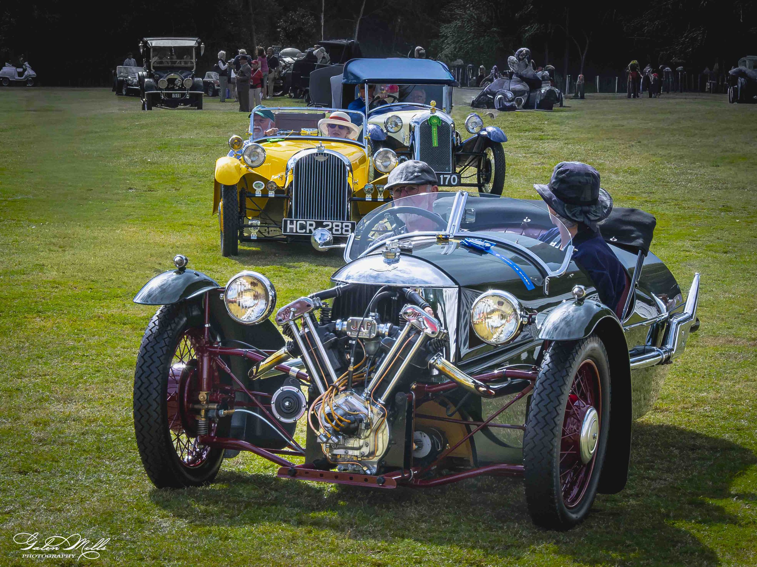 Vintage cars displayed outdoors at a classic car show with people viewing and chatting in the background.