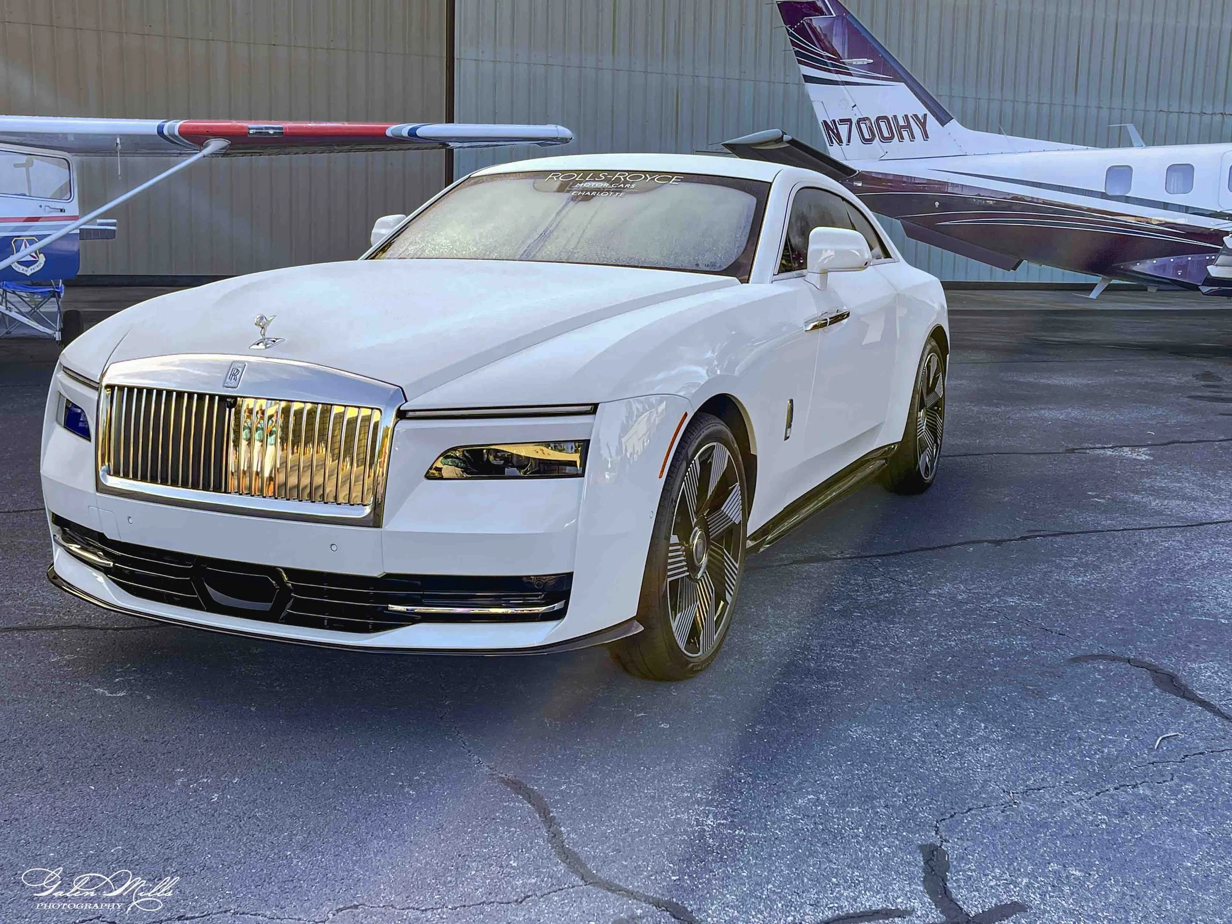 A white Rolls Royce car parked on a paved surface with small cracks, next to a small aircraft and a private jet, both parked outside a hangar with corrugated metal walls.