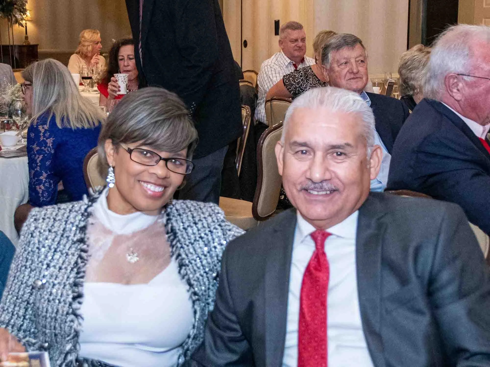 Smiling couple seated at a formal event, with people in the background in a banquet setting.
