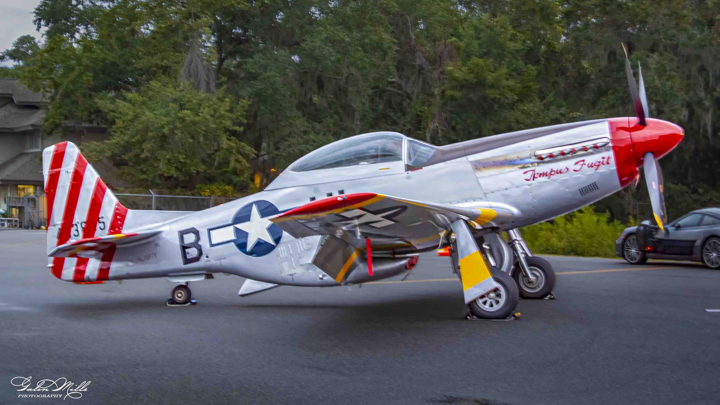 A vintage silver airplane with red and white striped tail and nose, parked on a tarmac with a car in the background and trees behind.